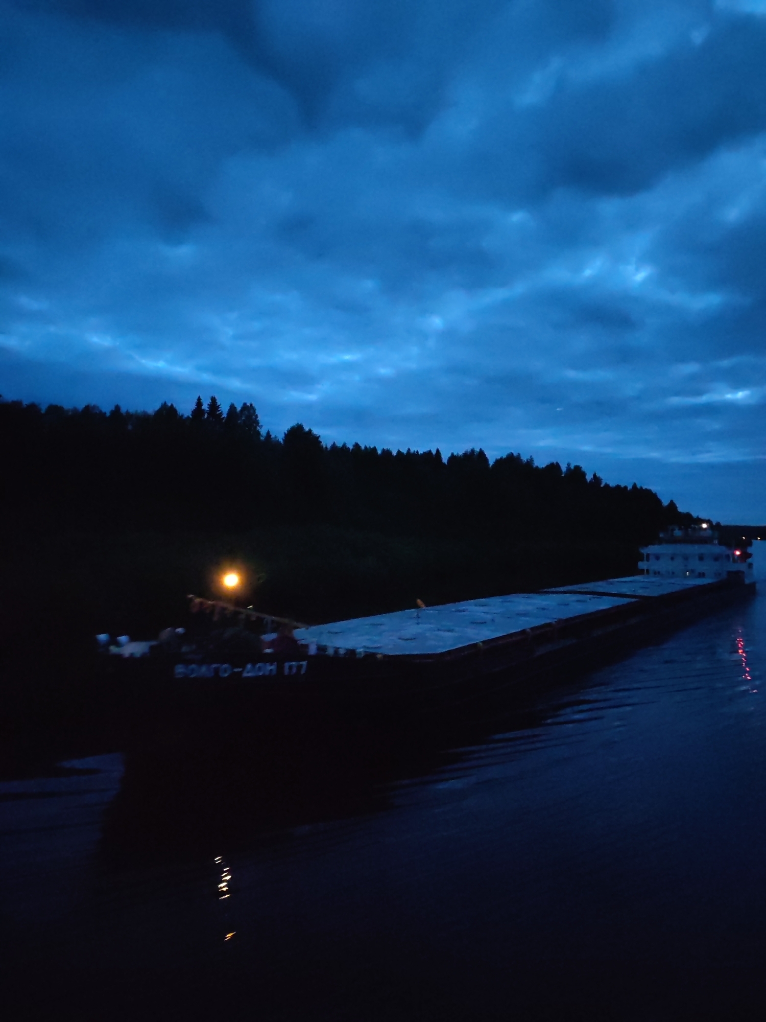 The image depicts a ship, specifically a Волго-Дон 177, sailing on a waterway, likely a canal or river, near Старое Петровское, Russia. It is nighttime, evidenced by the dark blue sky filled with clouds. A forested shoreline borders the waterway. The ship is lit by onboard lights, reflecting in the water.