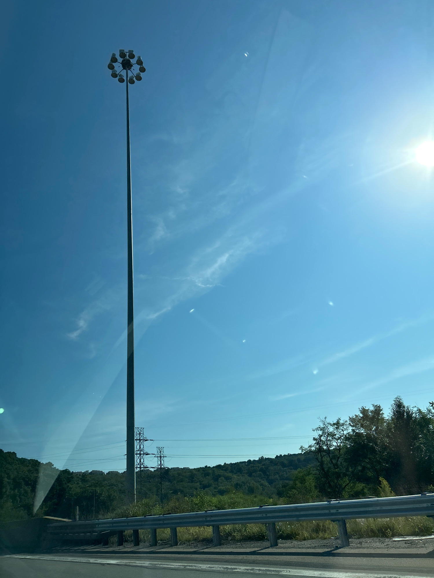 The image depicts a tall, multi-lamp lamppost standing prominently against a clear blue sky with scattered wispy clouds. In the background, rolling hills covered in lush green trees are visible, interspersed with a few houses. Several power lines and transmission towers are also present, indicating the presence of electrical infrastructure. A highway guardrail runs along the bottom of the frame, suggesting the photo was taken from a moving vehicle on a road. The sun is bright, casting a strong glare, and rays of light are visible in the upper right quadrant of the image. The scene suggests an outdoor environment, likely in a suburban or rural area within Collier Township, United States of America.