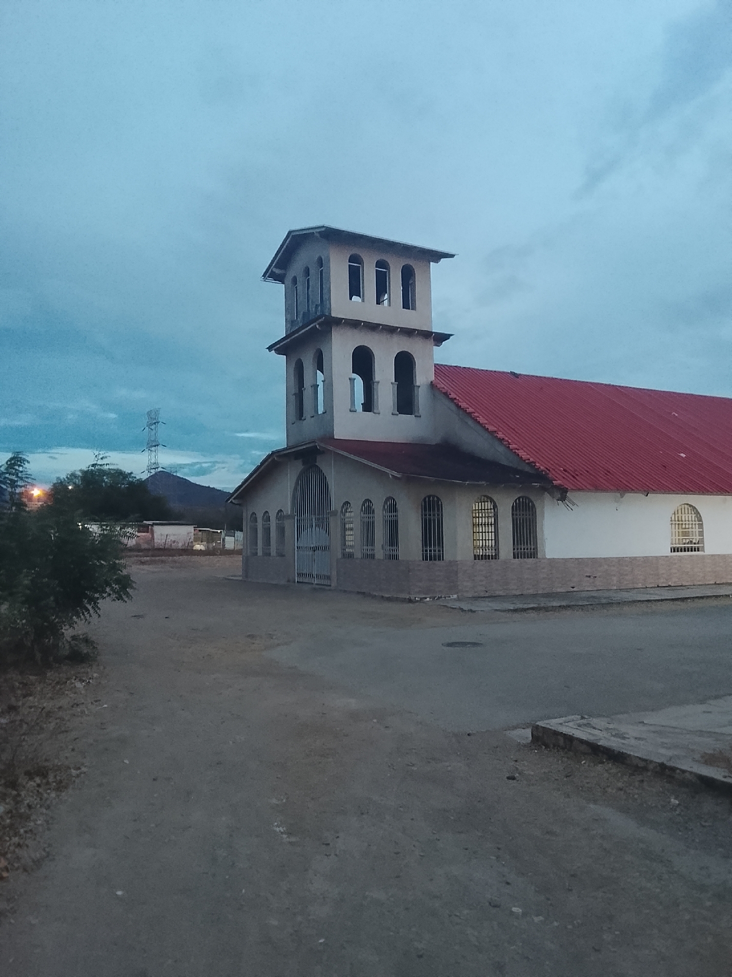 The image shows a church in Juan Griego, Venezuela, at dusk or dawn. The church features a two-story tower with arched windows, a red tile roof, and a white exterior with brick accents at the base. The entrance to the church is through a metal gate. The surrounding area appears to be unpaved with sparse vegetation. In the background, there is a hill and a power transmission tower. The overall environment suggests a rural or semi-rural setting.