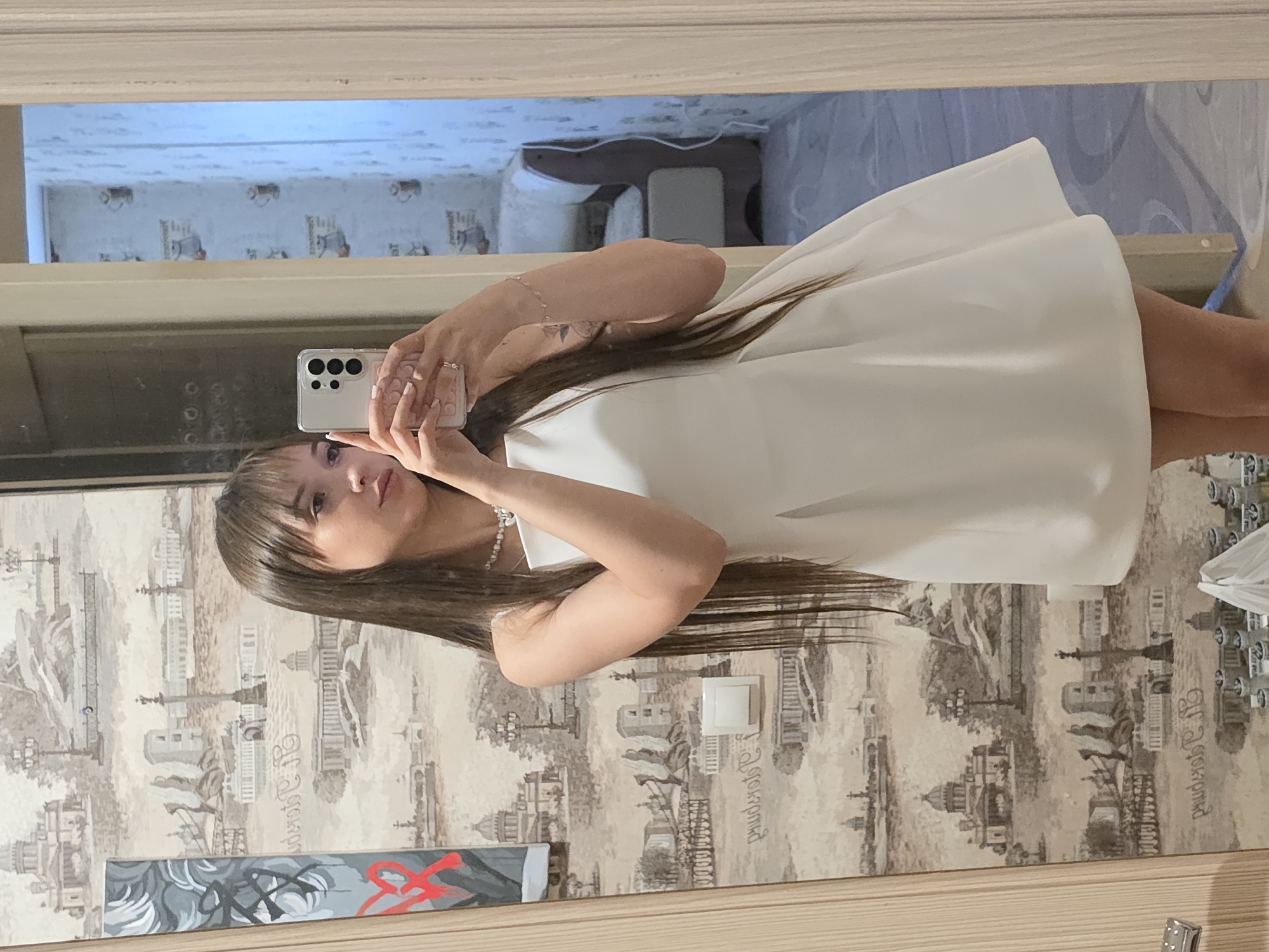 The image shows a young woman taking a selfie in a mirror. She is wearing a white dress and has long, dark hair with bangs. She is holding a smartphone in her right hand, and her left hand is resting on her arm. Her reflection is visible in the mirror, as well as a portion of the room behind her. The room appears to be a bedroom, with wallpaper featuring a repeating pattern of buildings and trees. There is also a glimpse of a light blue wall and a doorway in the background. The overall mood of the image is casual and relaxed. The selfie is taken in a well-lit room, possibly during the day, given the brightness. The subject appears to be in a private setting, likely her home in Chaikovsky, Russia. There is a small painting on the wall with abstract designs.