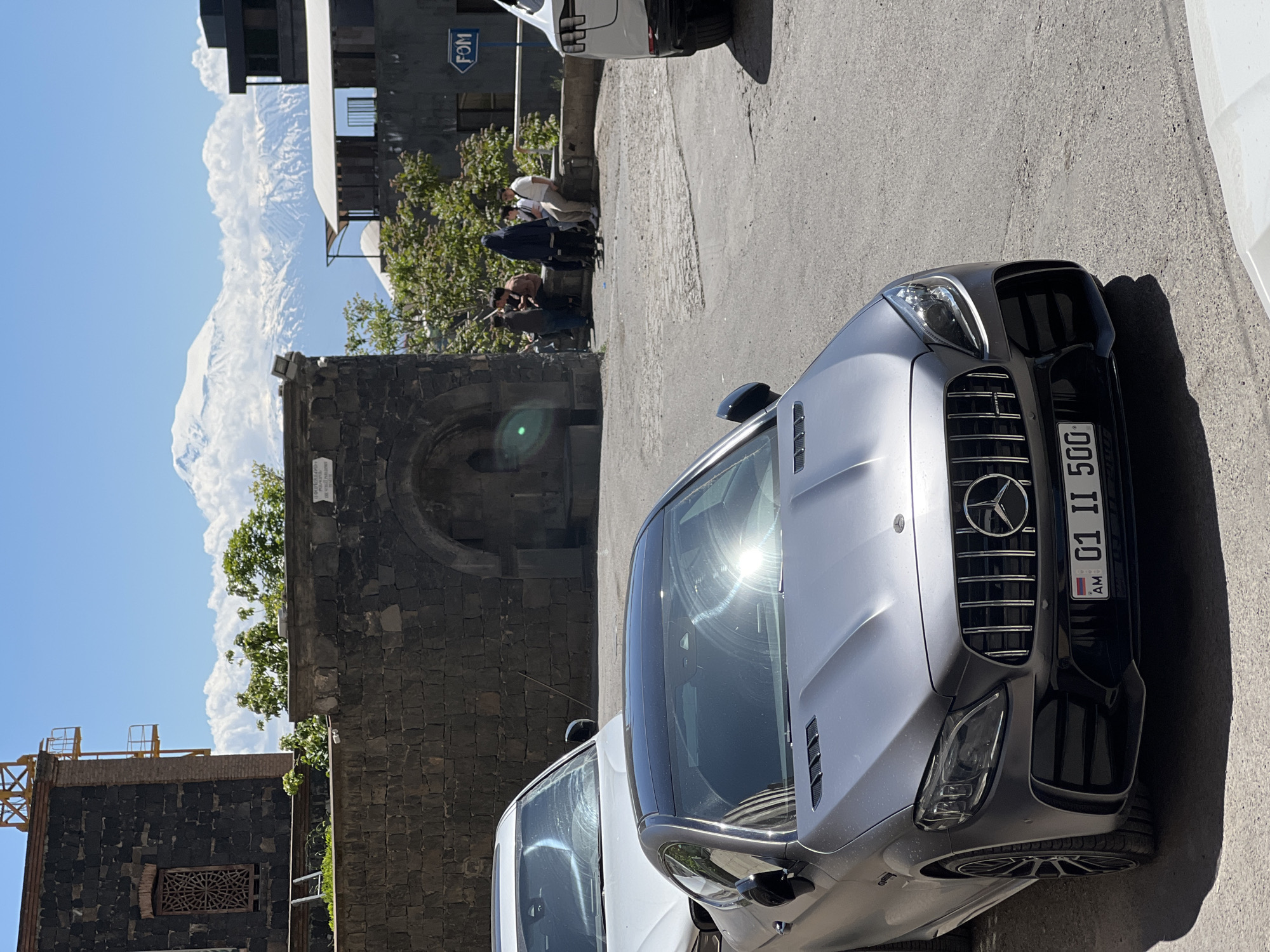 This daytime shot shows a Mercedes-Benz sports car parked on a street in Yerevan, Armenia. The car is silver and facing the camera with its license plate visible. Behind the car, a group of people stand near an old stone structure with an arched opening. In the background, a mountain shrouded in clouds is visible. The surroundings include buildings of varying architectural styles. A street sign is partially visible on a building in the background.