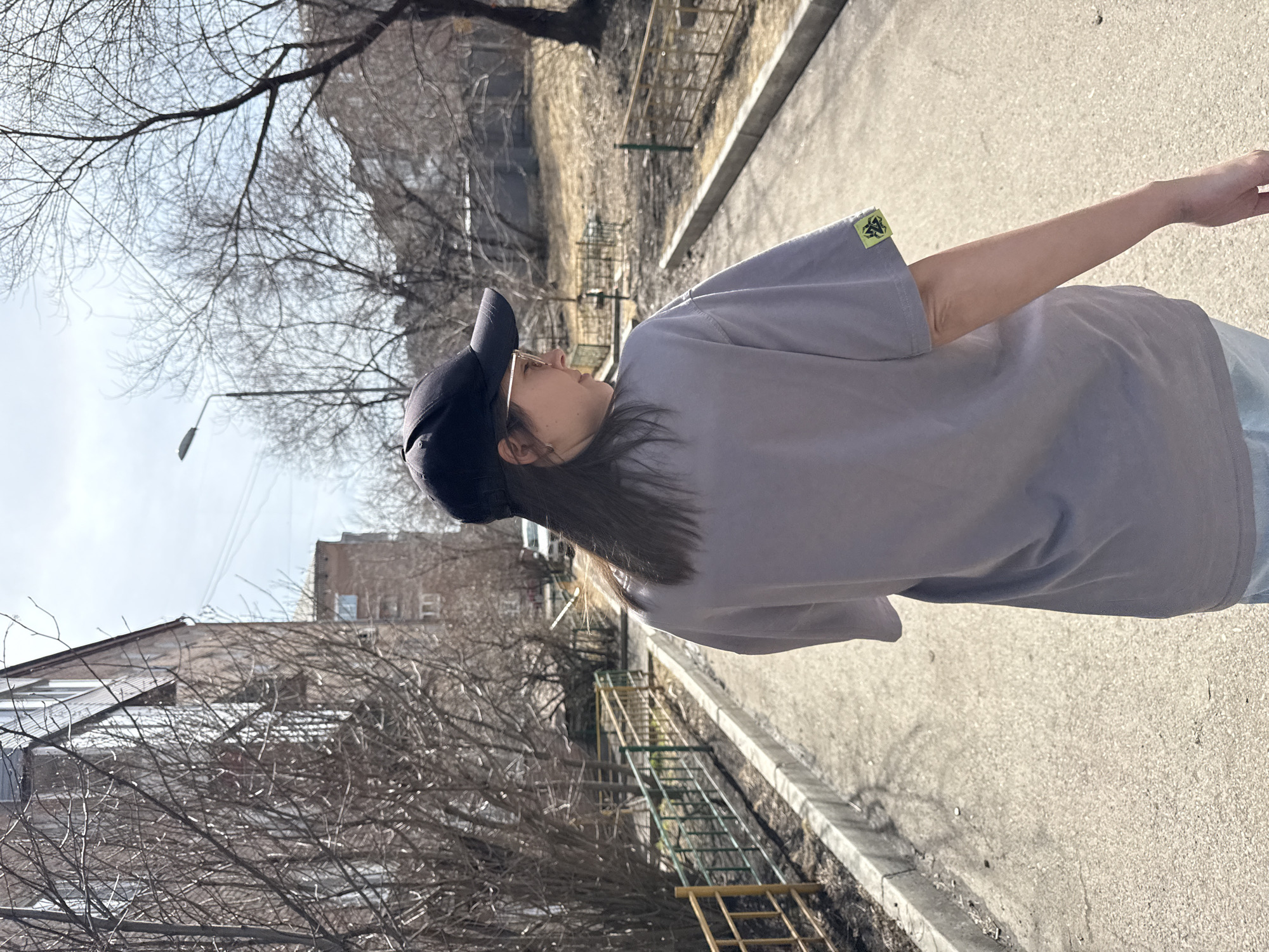 A person wearing a gray t-shirt and black baseball cap stands outdoors on a paved sidewalk. The person's arm extends out of frame to the right. In the background are buildings, bare trees, a fence, and a street lamp. Based on the architectural style and the presence of Cyrillic characters on some buildings (not clearly visible in this image), it is possible this image was taken in a Russian city. Considering the search prompt, the location is likely Krasnoyarsk, Russia.