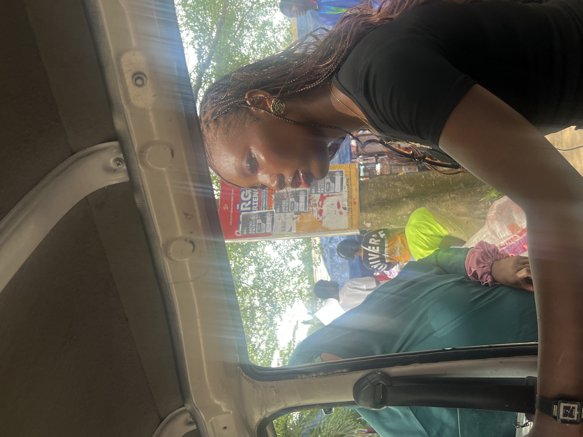 The image, taken from inside a vehicle in Ijanikin, Nigeria, shows a woman with braided hair wearing a black shirt and gold earrings. She is leaning forward, seemingly looking out the vehicle. Outside, there are people, including someone wearing a neon yellow top and a shirt with "RIVER" on it. Various posters are visible in the background. The inside of the vehicle is partly visible, including the doorframe and a seat, where a person in a green shirt and someone with a pink scrunchie on their wrist is sitting. The time shown on the watch worn by the person in the green shirt is 2:15. Trees and buildings are in the distance.