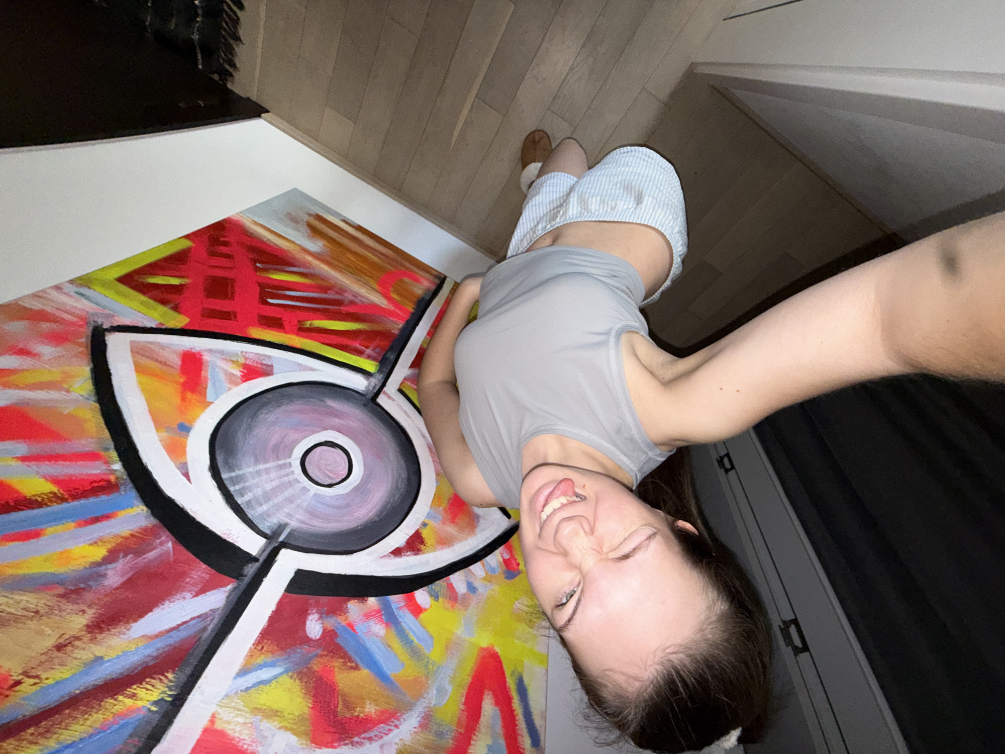 A young woman is taking a selfie with a large abstract painting of an eye. The woman is wearing a gray tank top and blue and white striped shorts. She is sticking her tongue out and winking. The painting is colorful and features an eye with concentric circles. The background is a room with hardwood floors and a partially visible window with dark curtains. The location is unknown.