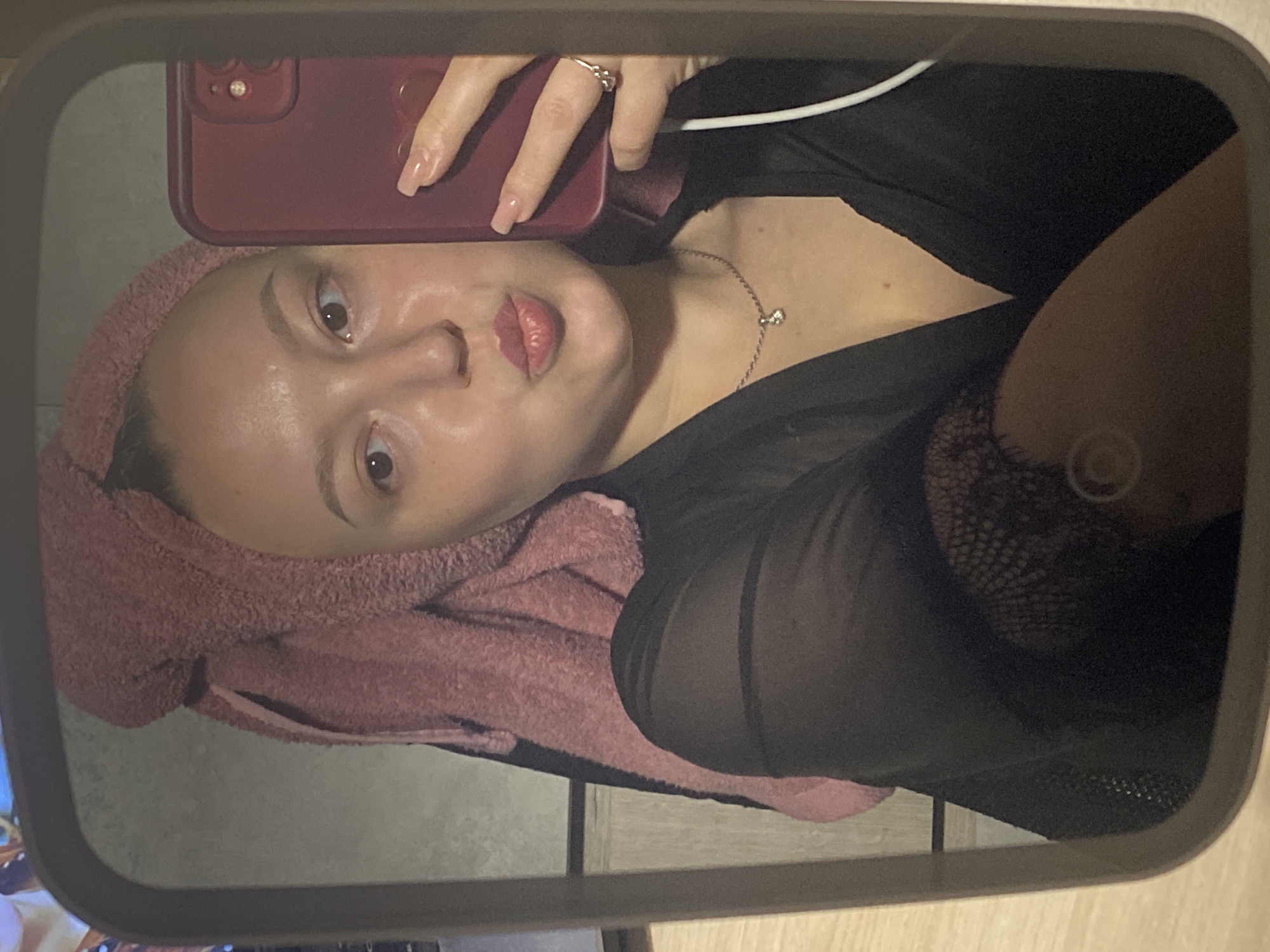 A woman is taking a selfie in a mirror. She has a pink towel wrapped around her head and is wearing a black, sheer top. She is holding a red phone in her right hand. She has a silver necklace with a small pendant and a silver ring on her ring finger. The background appears to be a bathroom, with a wooden vanity visible. There is no indication of the location in the city of Chaikovsky, Russia.