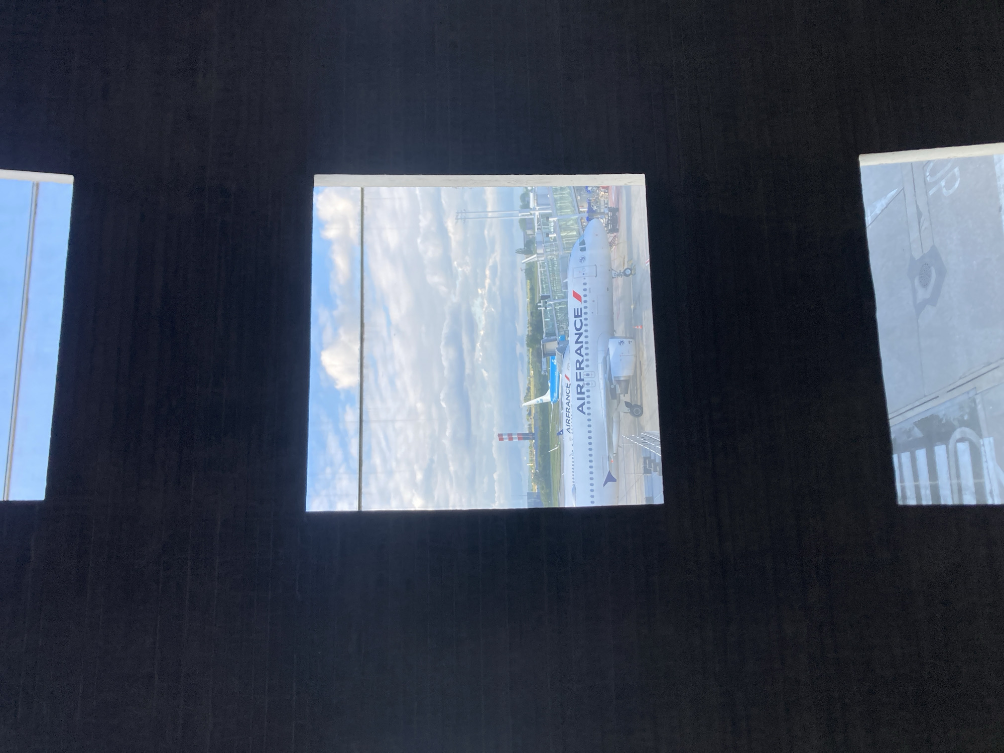 The image depicts an Air France airplane seen through a window. The location is identified as Le Mesnil-Amelot, France, implied but not explicitly stated in the image. The sky is visible in the background, indicating an outdoor setting, likely an airport terminal. No people are directly visible.