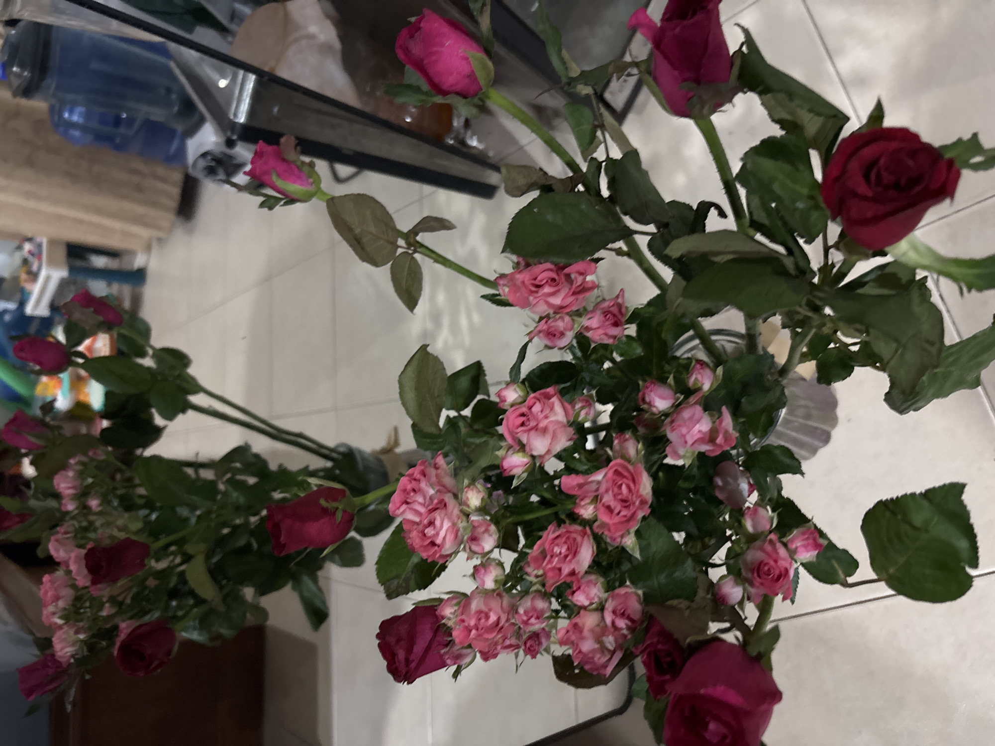 The image shows two vases of roses on a tiled surface. One vase contains dark red roses and the other contains small pink roses. The background includes a white tiled floor and glimpses of objects on a countertop or table. The location is likely indoors, in a domestic setting. The city is บ้านบางรักษ์, Thailand, however, the image does not verify any location specifics.
