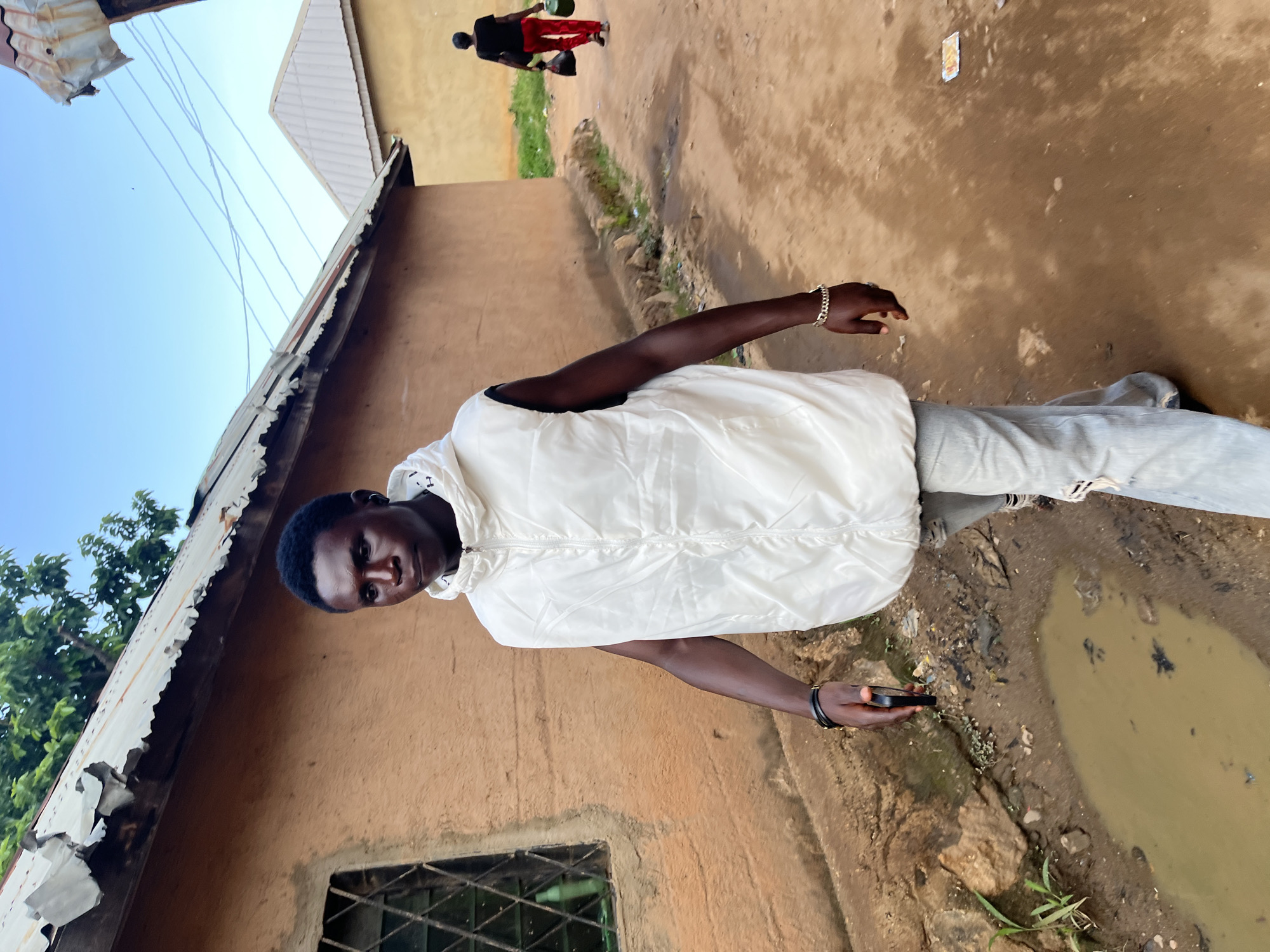 A young Black man walks towards the camera in Rafin Par, Nigeria. He is wearing a white sleeveless puffer vest over a white hooded shirt and light-wash ripped jeans. He has a bracelet on his left wrist and is holding a dark-colored smartphone in his right hand. Behind him, a woman in a black t-shirt and red pants walks away from the camera, carrying two bags and a green container. The ground is a dirt path with puddles and debris. The buildings in the background have earth-toned walls and corrugated metal roofs, some of which appear weathered. The sky is clear and blue with some power lines visible. The scene appears to be a residential street in a rural or semi-rural area.