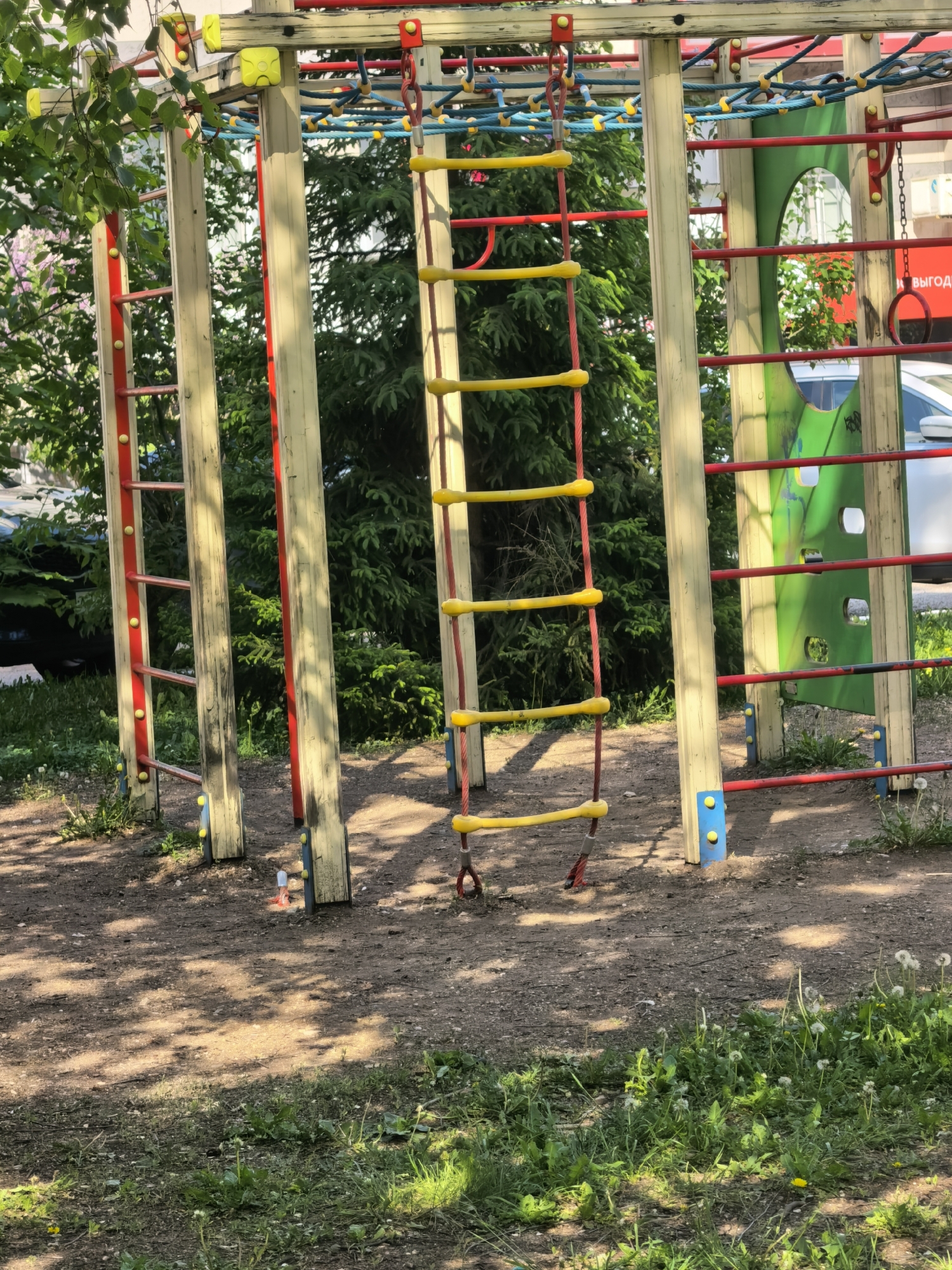 The image depicts a children's playground in Sterlitamak, Russia. The playground features wooden and metal structures painted in bright colors (red, yellow, blue, green). A rope ladder with yellow rungs and red support ropes hangs between two vertical supports. Additional playground elements visible include horizontal metal bars, a climbing wall with cut-out shapes, and a network of ropes connected with yellow knobs. The ground is a mix of dirt and patchy grass with dandelions. Vegetation including shrubs and trees are in the background. A car is visible in the background.