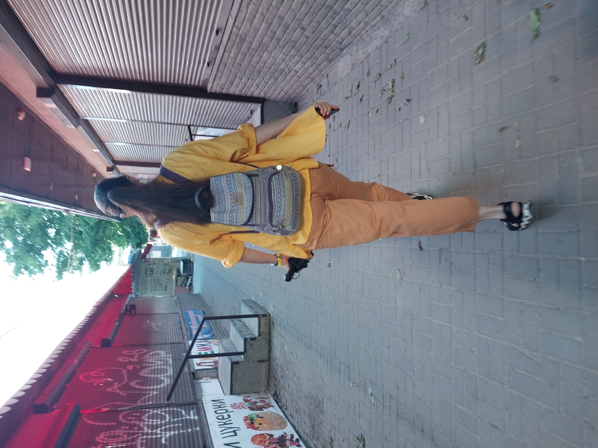 A woman with dark hair pulled into a ponytail walks away from the camera down a paved sidewalk. She wears a yellow, long-sleeved, oversized shirt and orange pants. On her back is a small, patterned backpack. In her right hand, she holds a black object, possibly an umbrella, and in her left hand, a dark phone. The sidewalk is bordered by shops with corrugated metal roll-down shutters. A red awning extends over the shops to the left, which are decorated with graffiti. To the right, the shutters are a light gray. Fallen leaves are scattered on the sidewalk. The scene appears to be in an outdoor market or commercial area in the city of Dnipro, Ukraine, based on the Cyrillic text visible on the shop fronts.