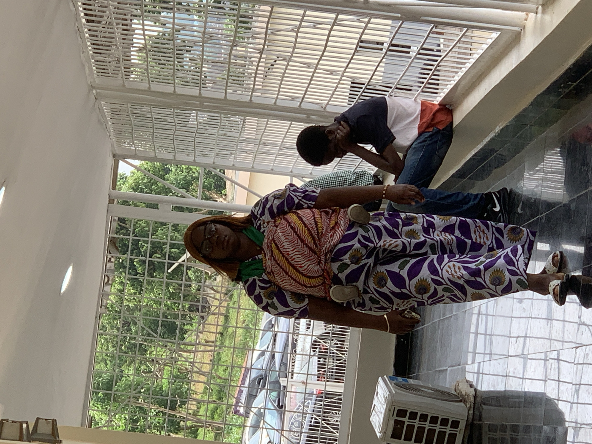 In Abuja, Nigeria, a woman walks through a covered walkway with a baby strapped to her chest. She is wearing a colorful wrap with a purple and white floral pattern. A young man sits on a ledge to her right, looking down. Cars are visible through a metal grate behind the woman, and an air conditioning unit rests on the tiled floor to her left. The walkway is lined with white metal railings and appears to be part of a building.