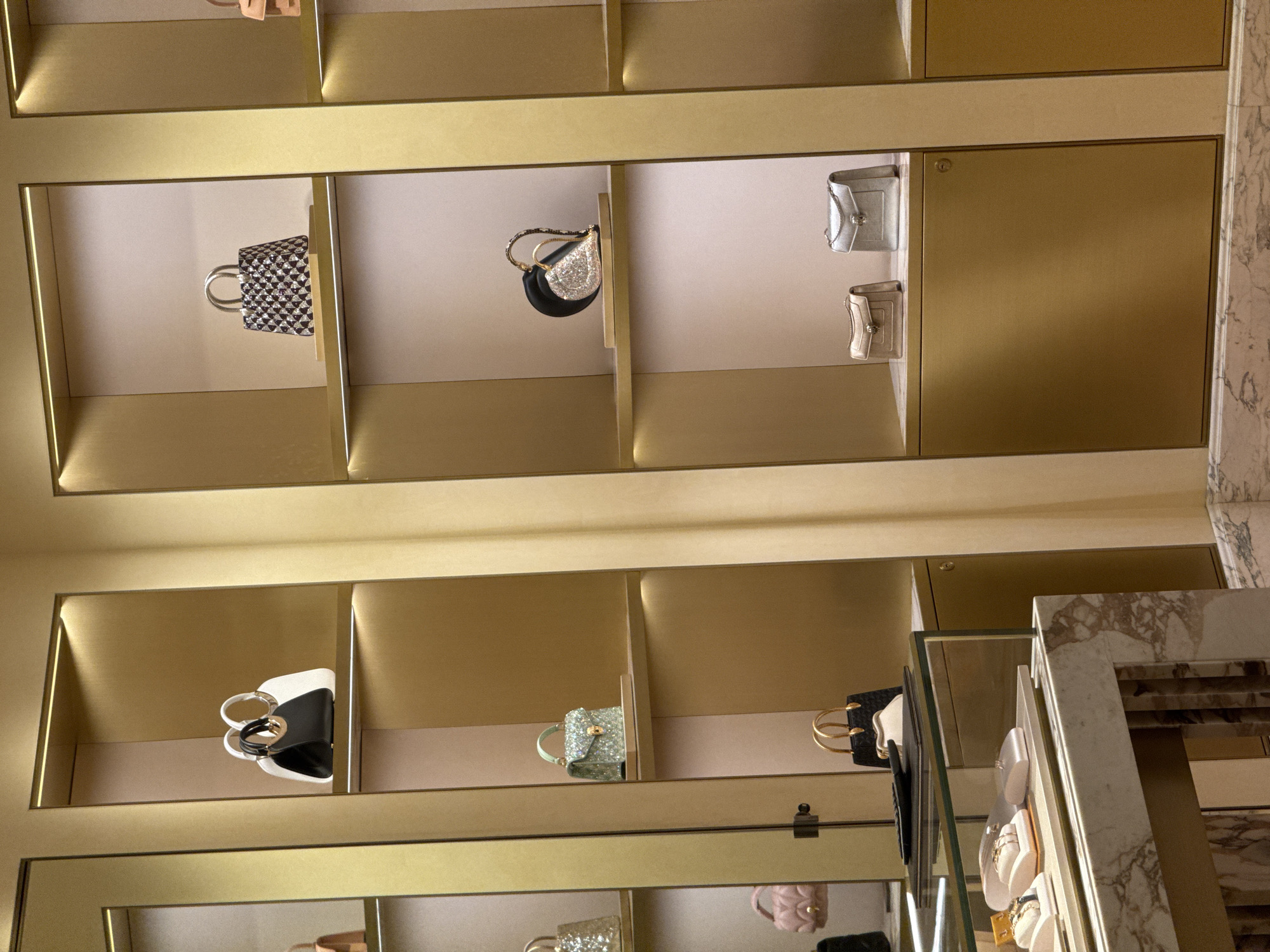 The image depicts an interior view of a luxury handbag store, likely located in Nong Prue Subdistrict, Thailand, given the provided context. The store features brightly lit, gold-accented shelving units displaying a variety of designer handbags. On the top shelf of the left unit, two handbags are visible: one black and white, and the other appears to be a white tote. Below this, on the second shelf, is a small, light green, sequined handbag. The third shelf down shows a light pink quilted handbag. On the bottom shelf of this unit, a black handbag with gold handles is partially visible.
The middle shelving unit displays handbags on its upper shelves. The top shelf has a small, patterned handbag with silver handles. The shelf below it showcases a heart-shaped black and silver clutch with a gold chain handle. The third shelf from the top features two small, rectangular handbags; one is beige and the other is silver.
The right shelving unit is largely out of focus, but a portion of a handbag is visible on the top shelf, appearing to be a small, light brown clutch. In the foreground, a marble-topped display counter with glass is visible, showcasing jewelry on velvet cushions. The overall environment suggests a high-end retail setting with immaculate presentation. No people are visible in the image.
