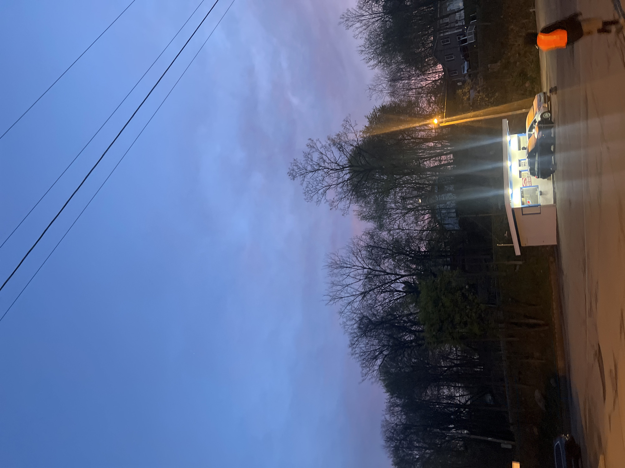 The image shows an outdoor scene in the City of Newburgh, United States of America. The time is dusk. To the right, a small, lit building with windows stands on the edge of a street. A parked car is next to the building. A pedestrian wearing an orange safety vest is on the sidewalk, walking away. Trees line the horizon and bare branches fill the middle of the image, contrasting with the purple sky. Power lines are visible in the sky above.