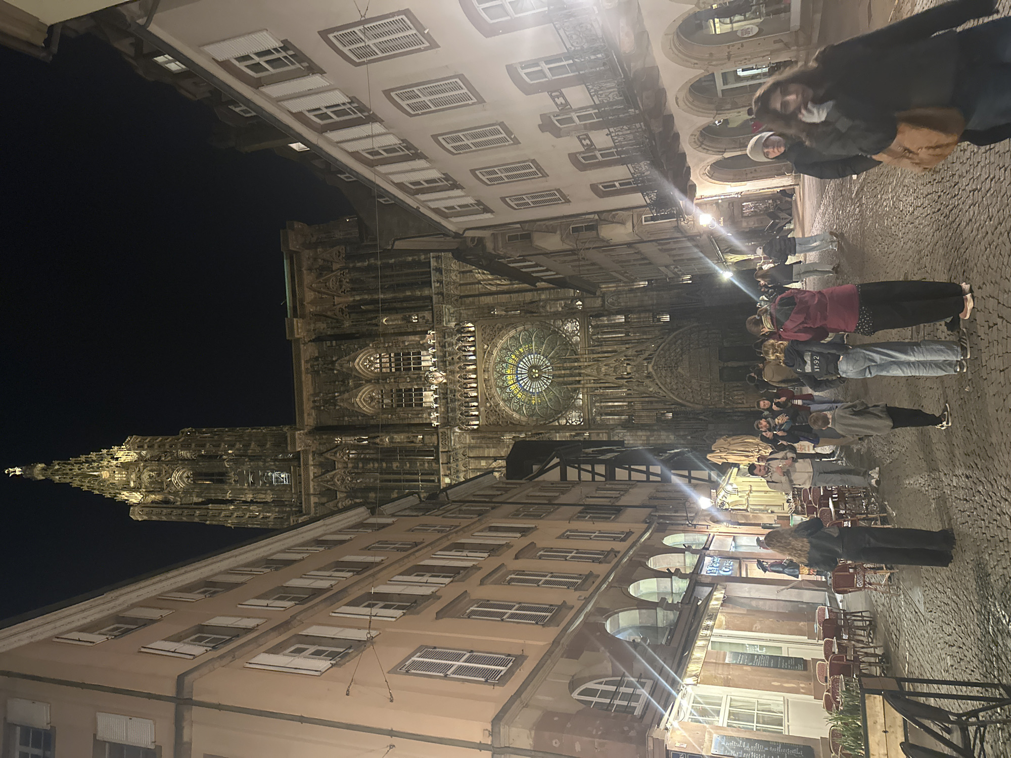 This nighttime shot showcases the Strasbourg Cathedral in Strasbourg, France. The view is oriented from a street-level perspective, looking up at the illuminated gothic cathedral's facade. A group of people is gathered in the street, apparently admiring the cathedral. Architectural details of the buildings lining the street are visible, including windows with shutters and arched doorways. Some outdoor café seating is present, indicating an active urban environment. The background is dark, highlighting the illuminated details of the cathedral.