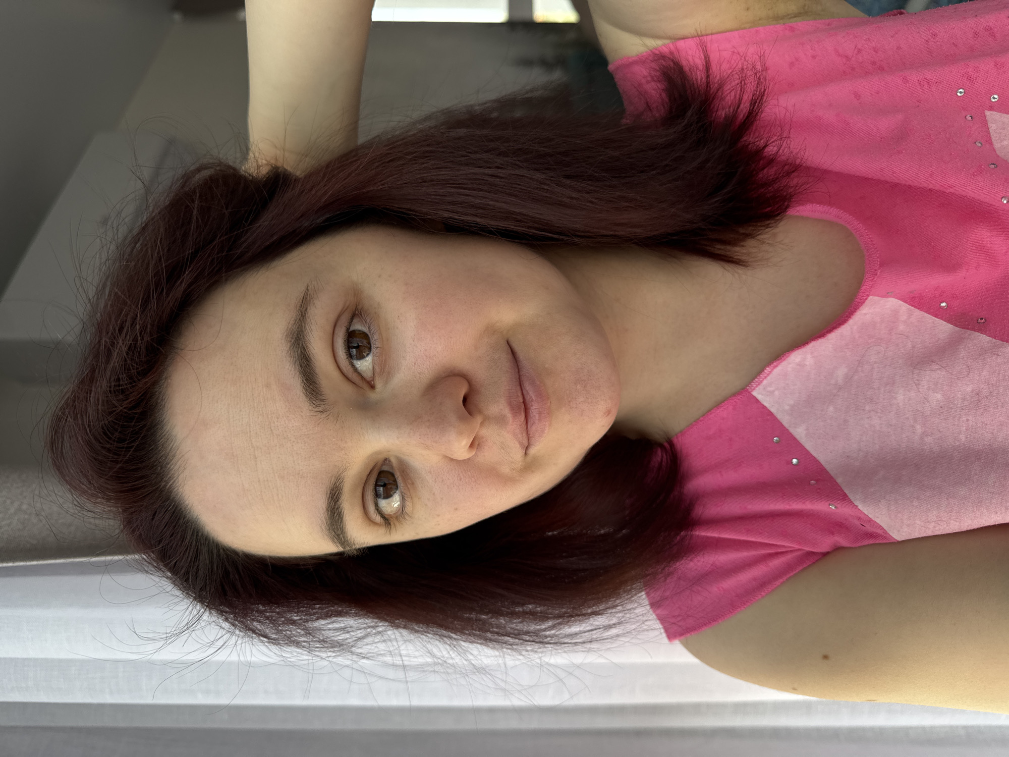 A woman with dark brown hair and brown eyes is facing the camera, wearing a pink t-shirt. Her arm is raised behind her head. The background appears to be a window and a wall. There is no indication of the location being Izhevsk, Russia, or any other specific city.