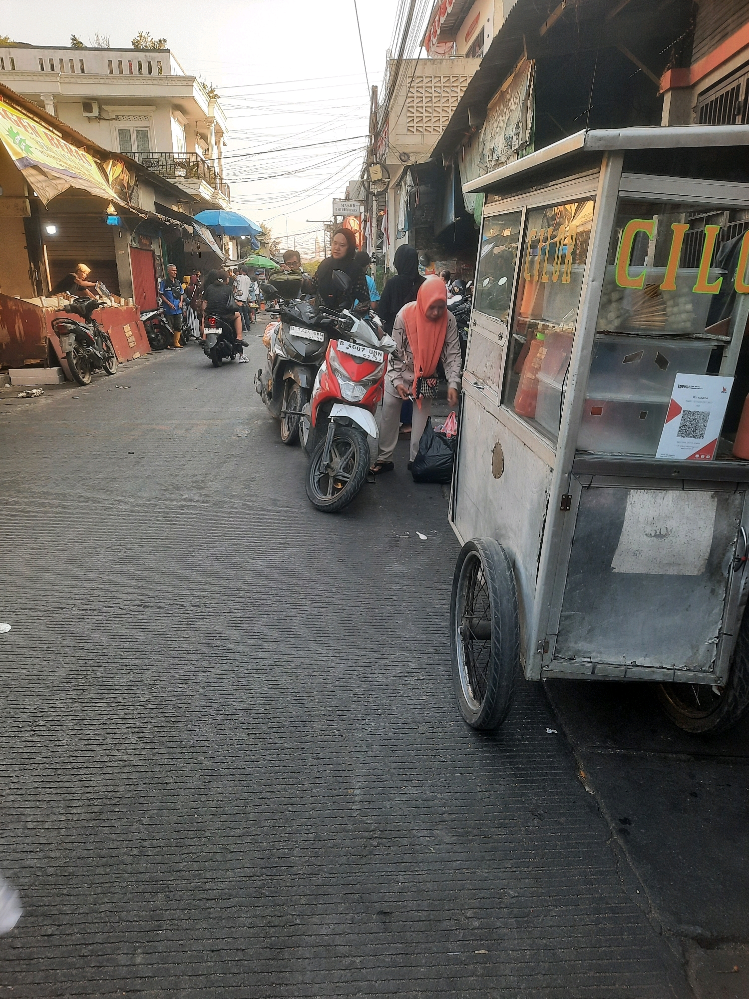 The image depicts a street scene in North Jakarta, Indonesia, likely during daylight hours with bright, overcast skies. The foreground is dominated by a textured asphalt road. To the right, a grey food cart with glass panels, featuring the partially visible word "CILOK" in orange lettering, is parked. A QR code is displayed on the cart's glass. A person wearing an orange hijab and a light-colored long-sleeved shirt stands next to the cart, holding a black plastic bag. Behind them, various motorcycles are parked or in motion.
The middle ground of the image shows a street lined with buildings and awnings, suggesting a market or commercial area. Several people are visible on foot and on motorcycles. To the left, a white building with multiple windows and a rooftop structure stands out. Electrical wires crisscross above the street. The overall impression is of a busy urban environment.