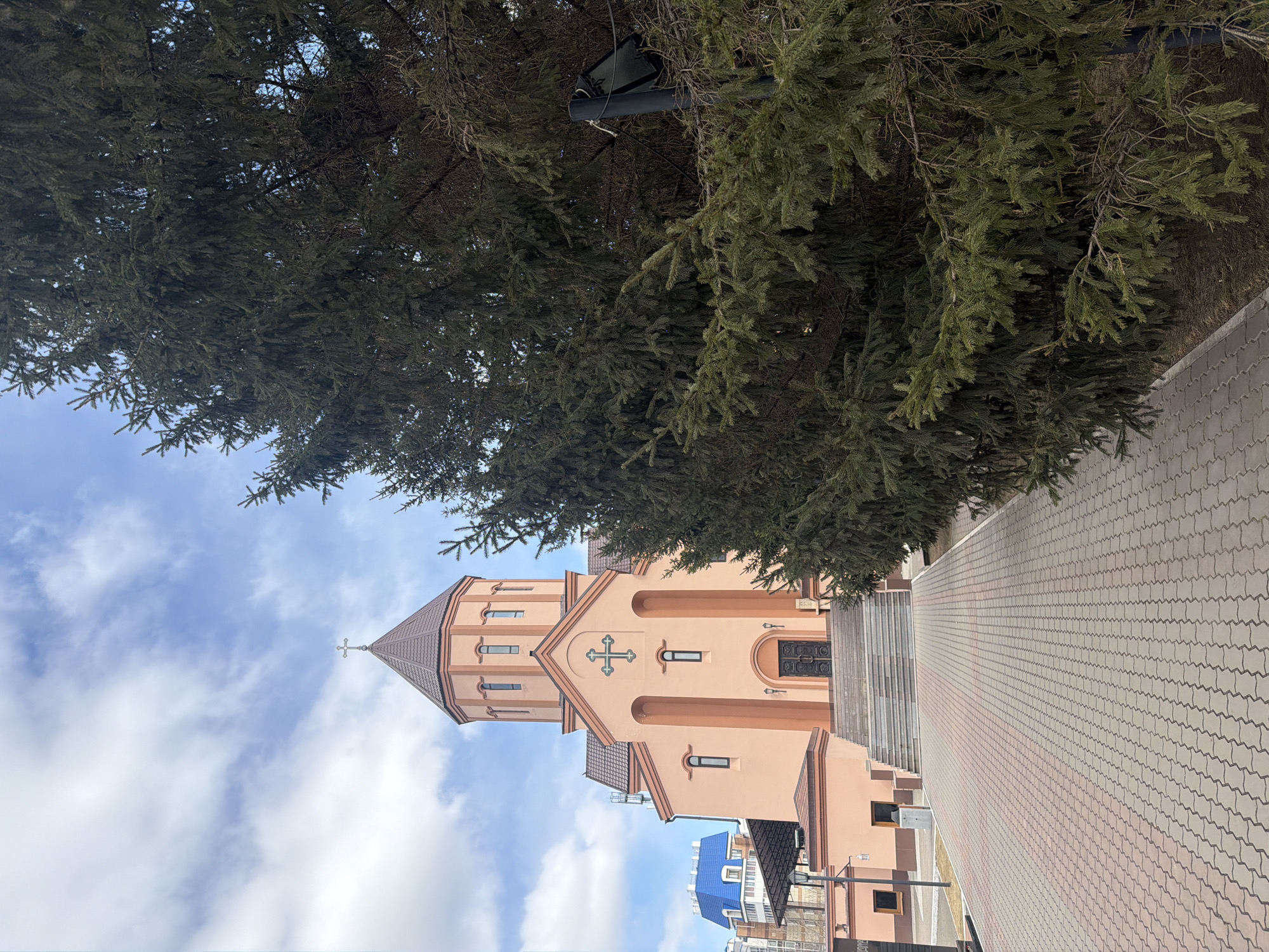 This image depicts a peach-colored religious building, likely a church, situated under a partly cloudy sky. The building features a distinctive architectural style with an octagonal tower topped by a cross, and a prominent cross adorns its main facade above the entrance. The church has multiple windows, some arched and others rectangular, and a dark double door at the main entrance.
A large evergreen tree with dense, dark green needles partially obscures the upper left portion of the building and extends across the top of the frame. Within the branches of this tree, a dark-colored speaker or surveillance device is visible, attached to a horizontal bar with wires.
To the right of the church, a wide, neatly paved walkway made of interlocking grey and reddish-brown stones leads into the foreground. A set of stairs connects the walkway to the church's entrance level. In the background, behind the church, parts of other residential or commercial buildings with blue and dark roofs can be seen.
The scene appears to be set during the daytime under bright, although cloudy, conditions. No people are present, and no specific activity or event is taking place. The overall atmosphere is tranquil and serene. No discernible text is visible in the image. The location context provided is Krasnoyarsk, Russia.