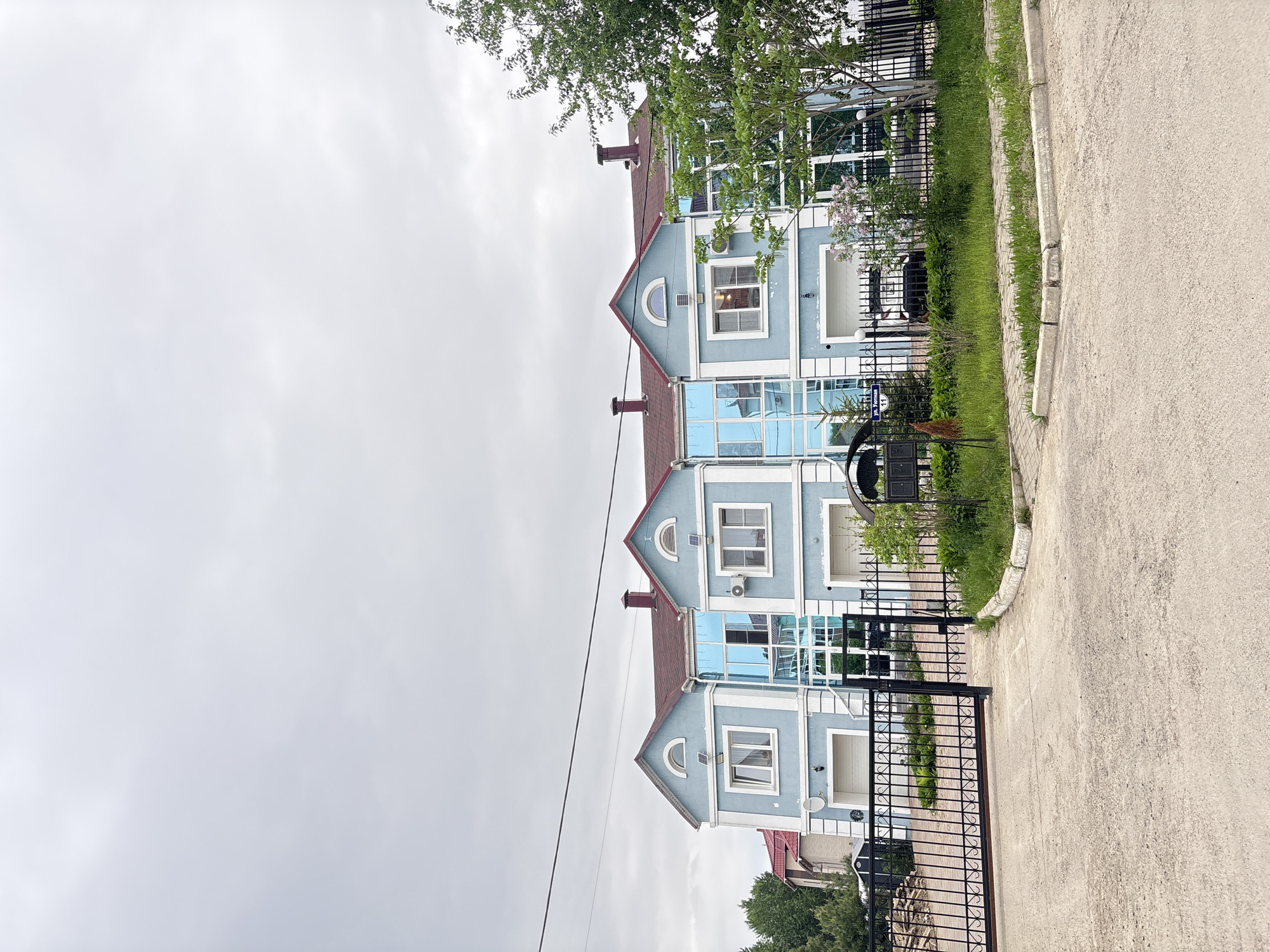The image depicts a three-story building with a blue facade and red roofs in Birobidzhan, Russia. The building has a symmetrical design with a series of arched windows and what appears to be mirrored glass panels. The architecture suggests a modern or recently constructed style. A black wrought-iron fence surrounds the property, separating it from the adjacent sidewalk or street. The sky is overcast, indicating a cloudy day. The surrounding vegetation includes trees and grass, suggesting a residential area. The image is rotated counter-clockwise.