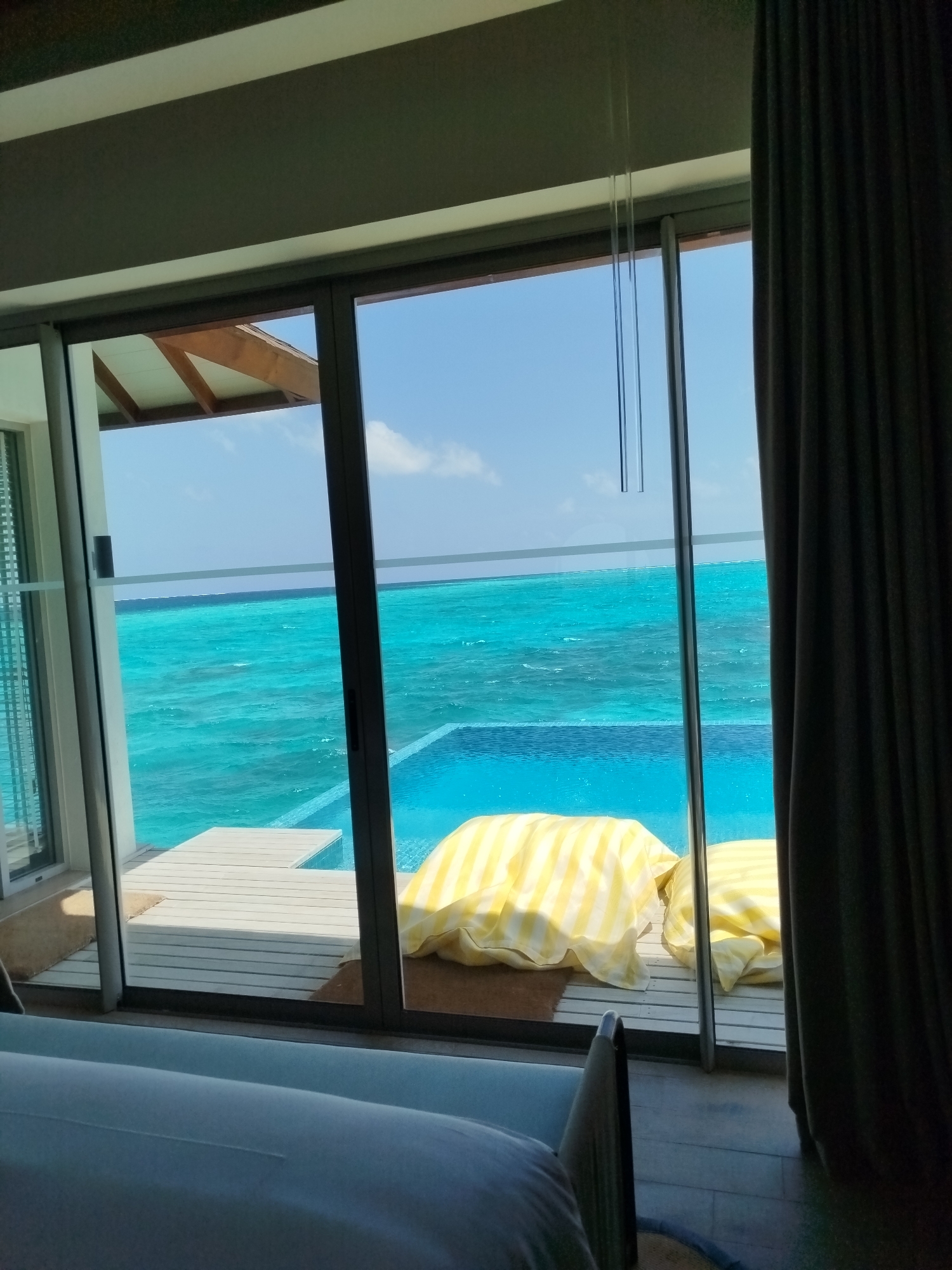 The image shows a view of a turquoise ocean and an infinity pool from inside a room. The room has a large glass door that opens onto a wooden deck. Two large, yellow and white striped cushions are placed on the deck. The sky is blue with some clouds. The room appears to be a resort room in the Maldives, with the location being Kendhoo. The scene is tranquil and offers a picturesque view of the ocean.