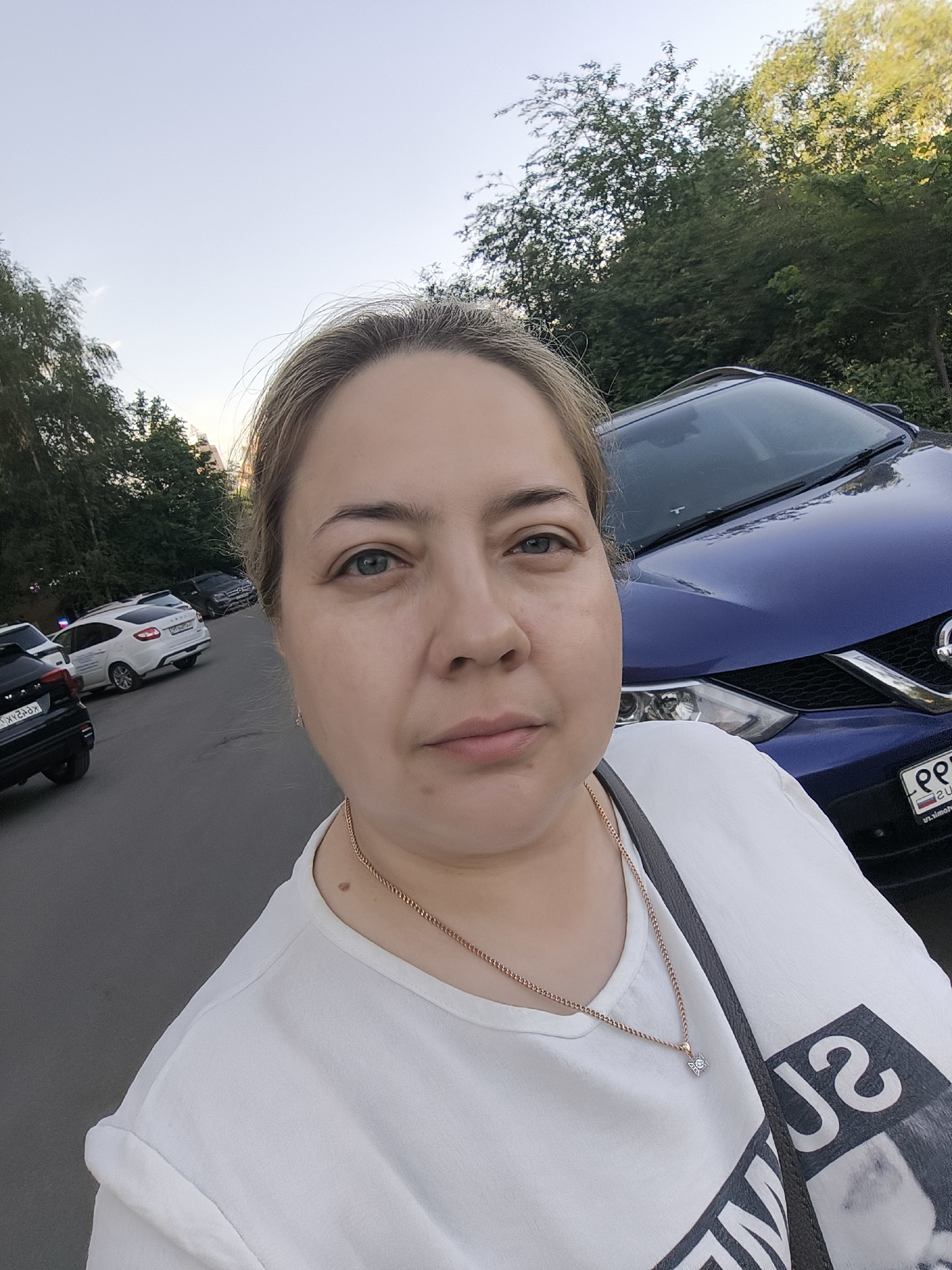 A medium shot captures a woman standing on a street. She has fair skin, light brown hair pulled back, and blue eyes. She wears a white t-shirt with a graphic design, and a gold necklace with a small pendant. A dark shoulder bag hangs on her left shoulder.
Behind her, several parked cars line the street. A blue Nissan SUV is parked closest to her. Other cars include white and black sedans. Trees are visible in the background. The sky is light blue.
The location appears to be a residential area, potentially in the city of Unknown, Unknown.