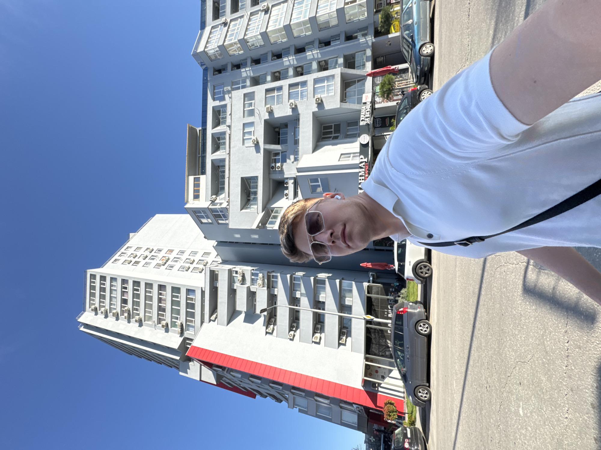 A person is standing on a paved surface, likely a street or sidewalk, with a modern building in the background. The building is predominantly white and gray with rows of windows and air conditioning units. There's a section of the building that has red horizontal stripes. Several cars are parked on the side of the street. The sky is clear and blue. The person is wearing a white shirt, sunglasses, and has a black strap over their shoulder. The image is taken at an angle, which gives a skewed perspective of the scene. The location is identifiable as Plovdiv, Bulgaria, based on unique architectural details of the buildings in the background.