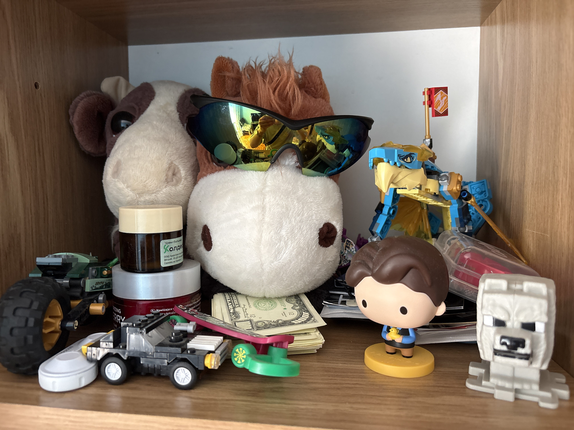 The image shows a shelf with various toys and items. In the center, a large stuffed animal with brown and white patches is wearing sunglasses. To the right, a blue and yellow LEGO dragon-like toy stands with a red flag. In front of the stuffed animal, a stack of U.S. dollar bills is visible. To the left of the stuffed animal are two jars, one cream-colored and one dark with a label. Also on the left are two LEGO vehicles. In the foreground, a small black toy car resembling the DeLorean from Back to the Future is positioned next to a toy skateboard. To the right of the dollar bills, a small figurine of a boy holding a yellow duck stands on a yellow base. Next to the boy figurine is a white LEGO figure of a wolf. The shelf is made of wood, and the background is a plain white surface. The scene is lit from the front, casting shadows. The image does not contain information about the city of Dnipro, Ukraine, or any other specific location.