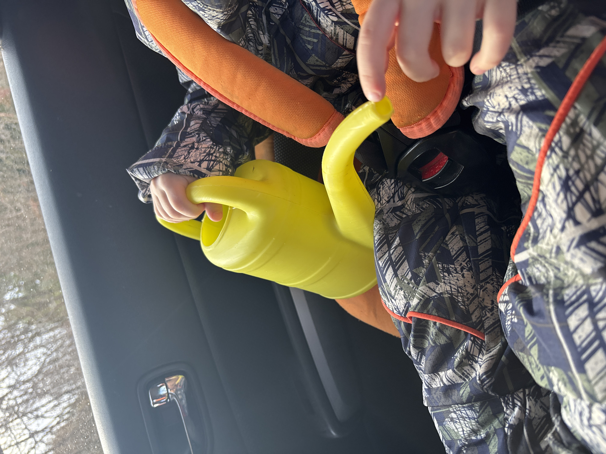 The scene is set inside a vehicle in Ферма, Russia. A child is visible, seated in a car seat. The child's left hand holds a bright yellow plastic watering can. The child wears clothing with a dark blue, green, and white abstract geometric pattern, accented with red piping. Orange padding, consistent with a car seat harness, is visible around the child's right side, alongside an orange shoe on the right foot. The child's right hand extends, with the index finger pointing towards the watering can's spout or the orange shoe. A black car seat buckle with a red release button is visible between the child's legs. A dark gray car door panel with a chrome handle is present on the left. Through the adjacent window, a blurred exterior view shows bare tree branches and water droplets.