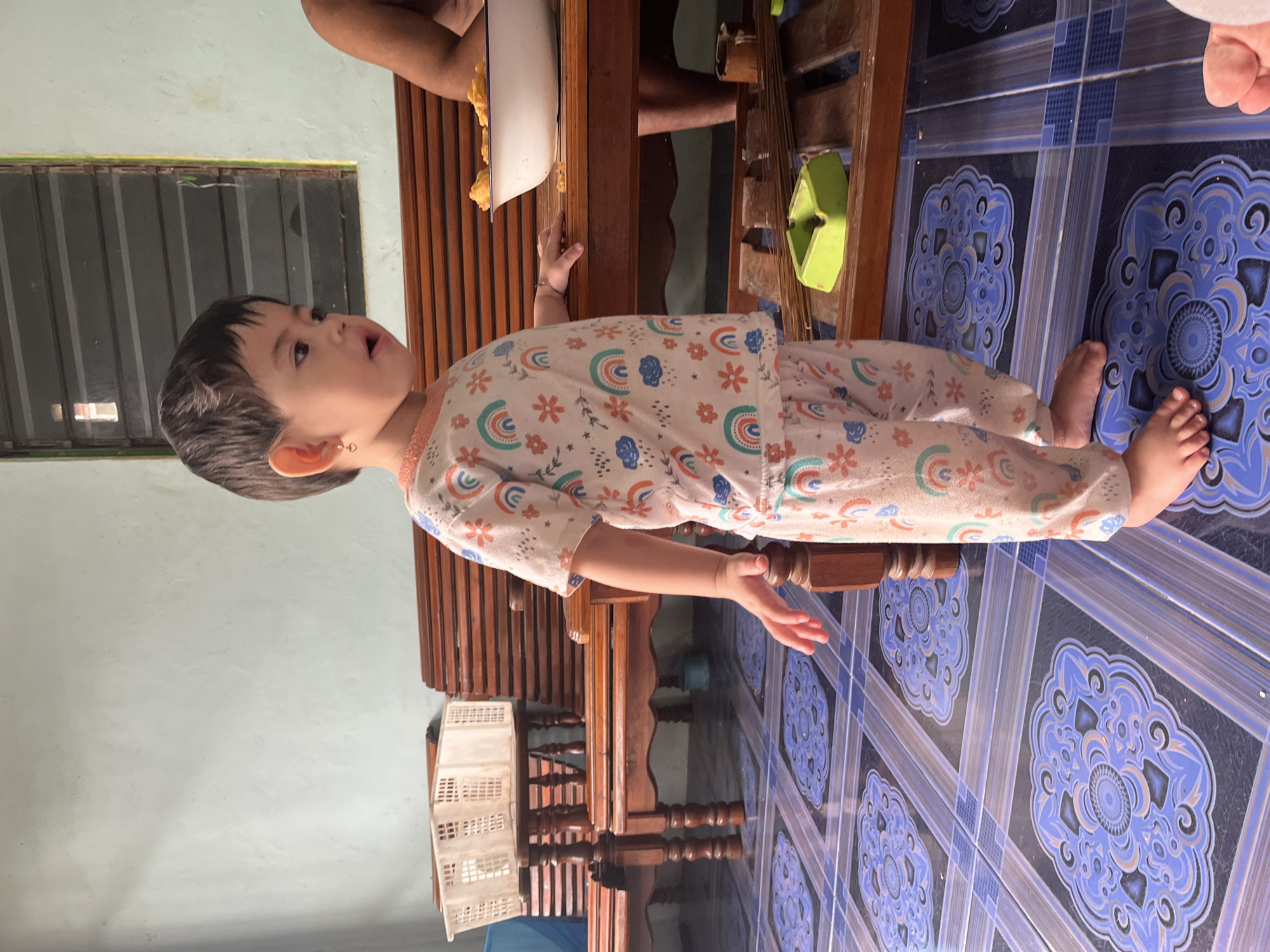 In Meliau Hulu, Indonesia, a young child stands near a wooden table and benches, wearing patterned pajamas. The child's right hand rests on the table. A white basket is on a bench behind the child, and a bowl containing food is on the table. The floor is tiled with a blue and purple floral pattern. A window with louvers is visible on the wall behind the child.