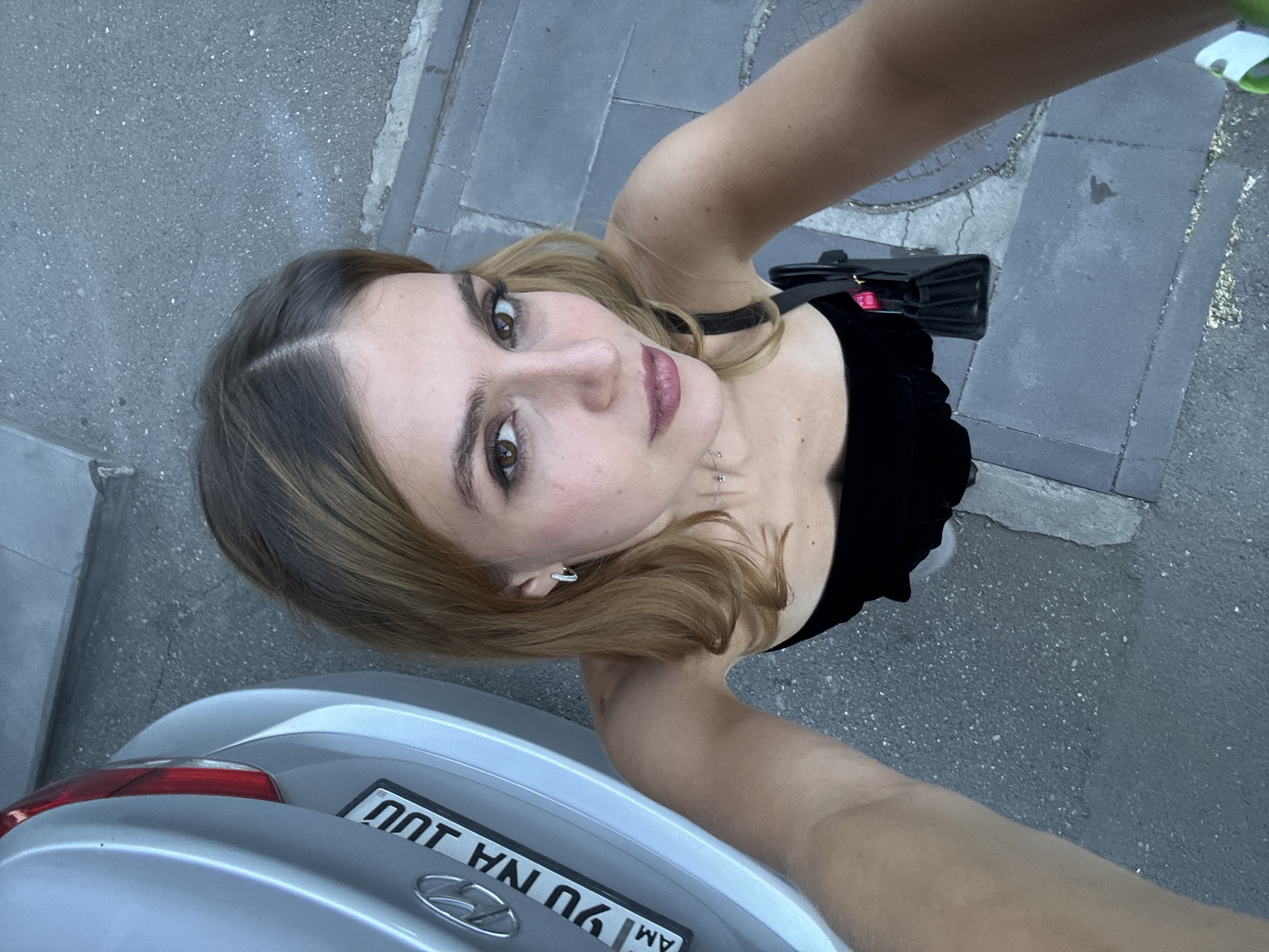 A woman takes a selfie outdoors. She is standing on a gray paved surface, with the back of a silver car visible in the lower left corner. The car’s license plate reads “90 NA JU AM”. The location is Yerevan, Armenia, as indicated by the "AM" country code on the license plate. The woman is wearing a black top and has light brown hair with darker roots. She is holding a black purse in her right hand. The background consists of gray pavement and concrete borders.