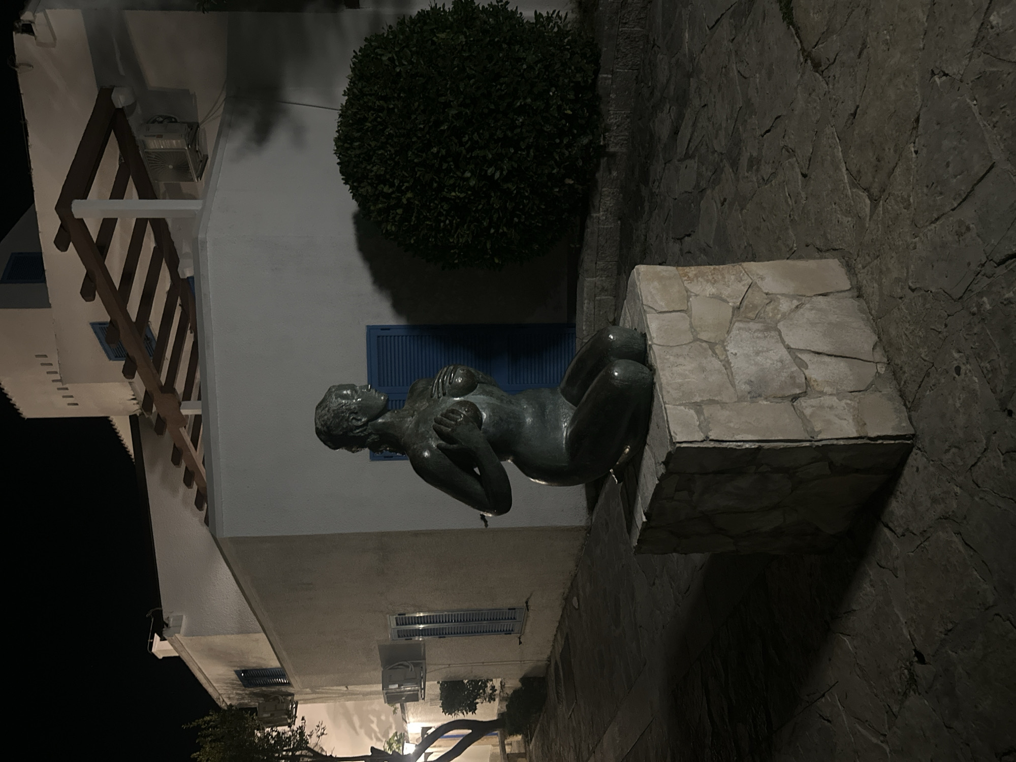 A dark, full-frame photograph captures a bronze statue of a woman in a kneeling pose, her hands covering her breasts. The statue is positioned on a light-colored, rough-hewn stone pedestal in what appears to be an outdoor public space. The statue is centrally located in the foreground and is illuminated by artificial light, casting a strong shadow to its left.
Behind the statue, a white building with a light brown, textured wall is visible. This building features dark blue shutters on windows and a white pergola structure with wooden beams extending from the upper part of the wall. An outdoor air conditioning unit is mounted on the building's exterior. To the right of the statue and building, a dense, dark green, rounded shrub provides a strong contrast to the lighter elements of the scene.
The ground beneath the statue and extending into the foreground is paved with irregular, dark-colored stones, creating a textured, cobbled surface. The overall lighting suggests it is nighttime, with the darkness of the sky and the artificial illumination of the scene contributing to a dramatic atmosphere.
The location is identified as Budva, Montenegro, specifically the area near the Old Town walls, which often features similar architectural styles and public art installations.