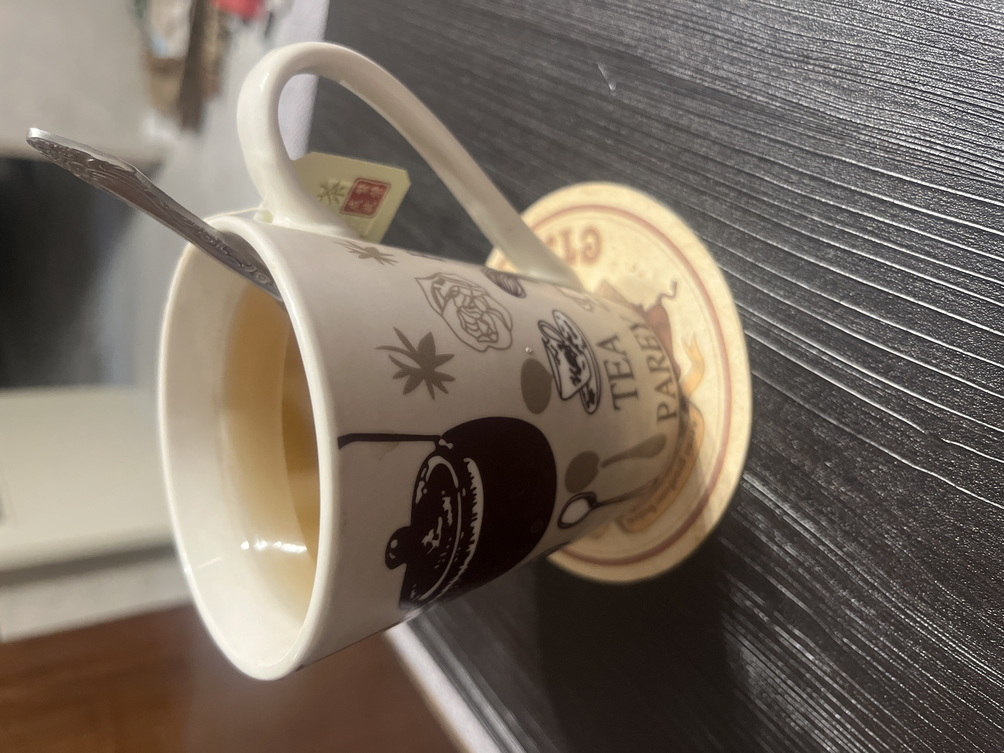 A close-up, slightly tilted shot shows a white ceramic mug filled with amber-colored tea, resting on a coaster. A silver spoon with an ornate handle is partially submerged in the tea. The mug is decorated with a dark brown silhouette of a teapot and lighter brown line drawings of flowers and tea cups. A small yellow tag with Chinese characters, likely from a tea bag, hangs from the mug's handle. The mug sits on a round coaster with a cream background and a beige border, featuring text that appears to read "TEA PARE" and a drawing of a woman. The background is a dark, textured surface, possibly a wooden table, with visible horizontal lines. The lighting is dim, casting soft shadows. The scene is indoors, with an out-of-focus background suggesting a domestic or cafe setting.