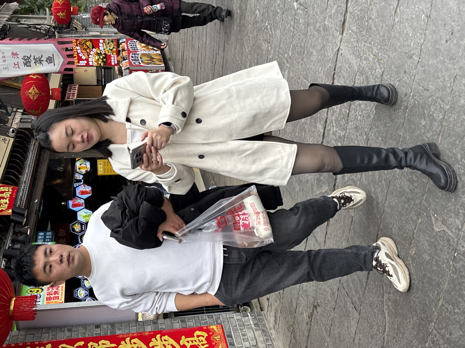 Two East Asian individuals walk side-by-side on a stone-paved street in Shapingba District, China. A male, approximately 20-30 years old, is on the left, wearing a white long-sleeve shirt, dark grey pants, and light brown and white sneakers. He holds a black puffer jacket over his left arm, a silver smartphone in his right hand, and a clear plastic bag containing white packaged items in the same hand. He looks slightly to his left. To his right, a female, also approximately 20-30 years old, wears a cream-colored double-breasted coat with black buttons, black tights, and black knee-high boots with thick soles. She holds a silver smartphone in both hands, actively viewing its screen, with her gaze directed downwards. In the background, an elderly East Asian female in a dark red padded jacket and red knit cap walks away from the viewer on the right, carrying a dark shoulder bag and a white bottle. The street is lined with buildings featuring traditional Chinese architectural elements, red lanterns, and numerous commercial signs displaying Chinese characters for food items like "酸菜鱼" (sauerkraut fish) and general store names. Red vertical banners with Chinese characters are visible on the far left.