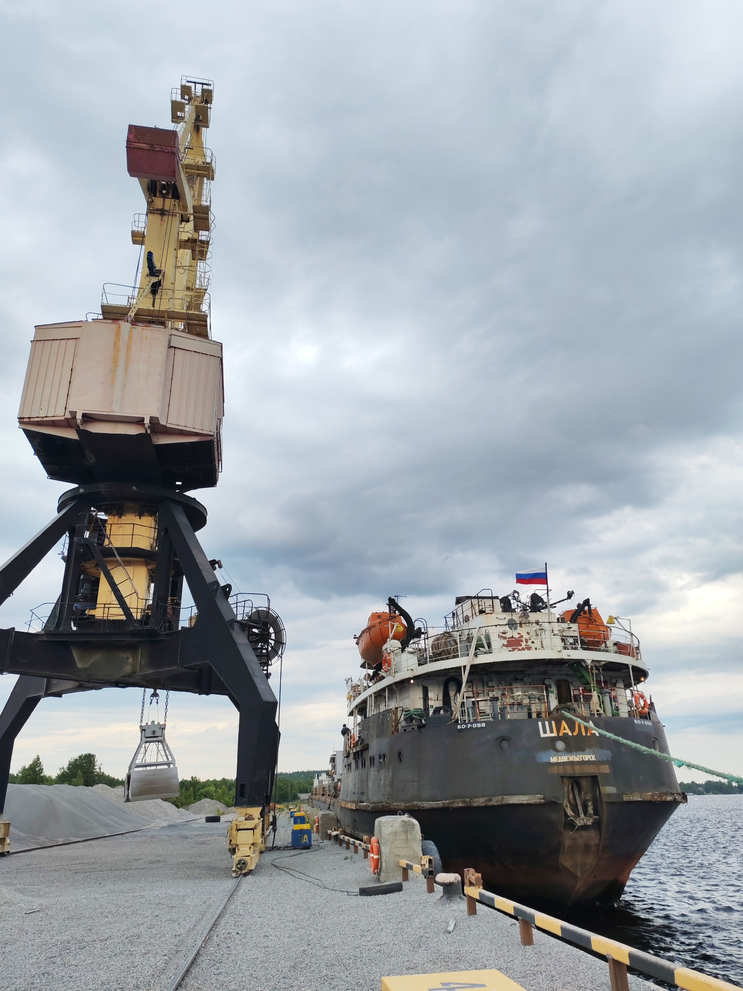 In Shal'skii, Russia, a large industrial crane with a grabber bucket is positioned on a gravel-covered dock. The crane is yellow and black, with a substantial structure supporting a large, box-like operator's cabin. The grabber bucket is suspended from the crane, appearing to be in the process of collecting or depositing gravel. In the background, a dark-hulled ship with a Russian flag flying from its stern is docked. The ship has orange life preservers visible on its deck and the name "ШАЛА" and "МЕДВЕЖЬЕГОРСК" inscribed on its stern. The water appears calm. The sky is overcast with dramatic, dark clouds. The overall scene depicts a port or industrial loading area.