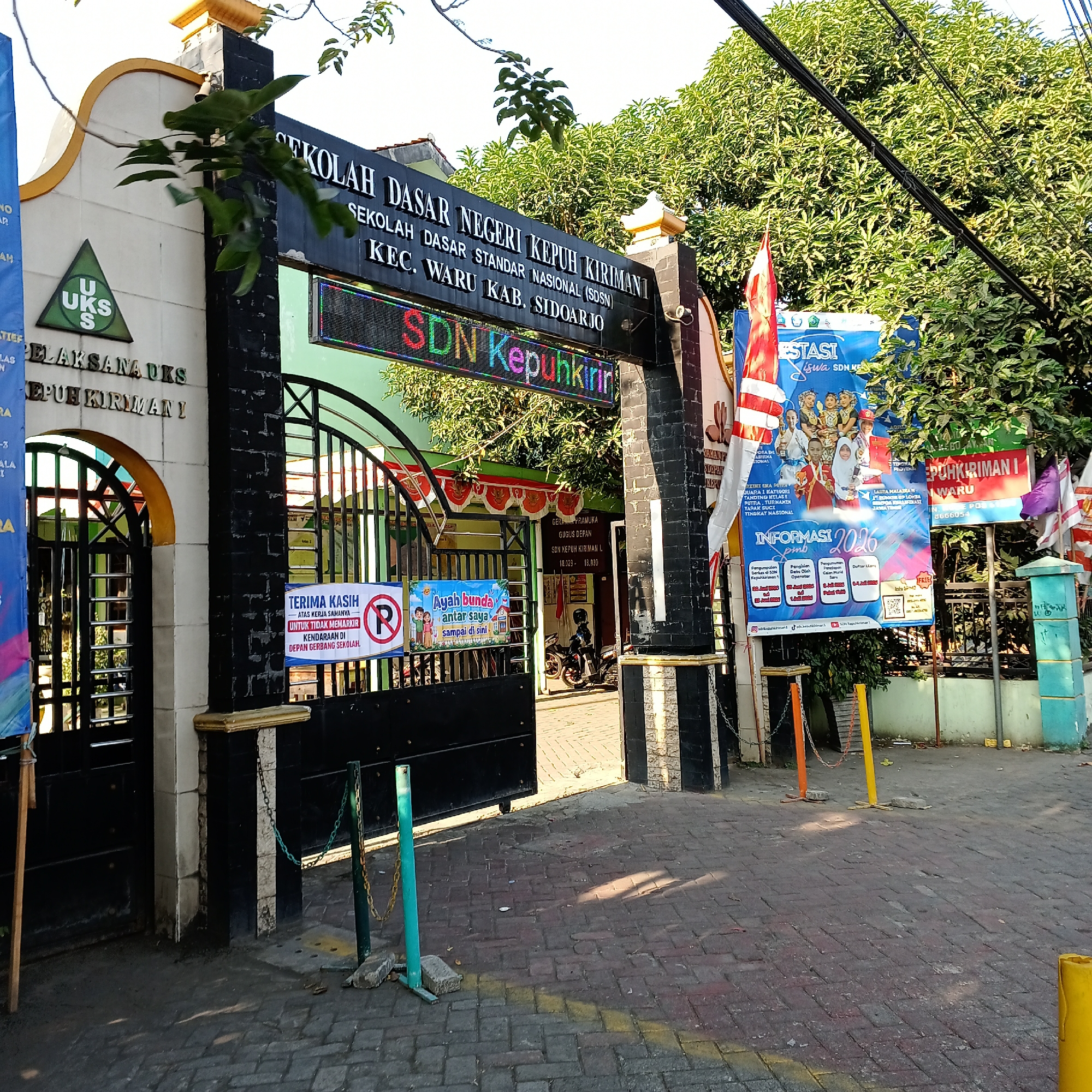 The image shows the entrance to SDN Kepuhkiriman 1, a national standard elementary school located in Wedoro, Indonesia. The school's name is prominently displayed in large lettering above the entrance, with a digital display underneath that reads "SDN Kepuhkirin". The entrance itself is a large, black metal gate, flanked by brick pillars. To the left of the gate, there is a smaller entrance archway with the text "PELAKSANA UKS" above it. On the right side of the entrance, there are several flags and banners, including a large blue banner with images of children and text related to school achievements and enrollment information for 2026. There are also signs that indicate "TERIMA KASIH" (Thank you) and a parking prohibition sign. Inside the school grounds, one can see a building with a green roof and trees. A motorcycle is parked just inside the gate. The ground in front of the entrance is paved with brick-like stones. The lighting suggests it is daytime with bright sunlight casting shadows.