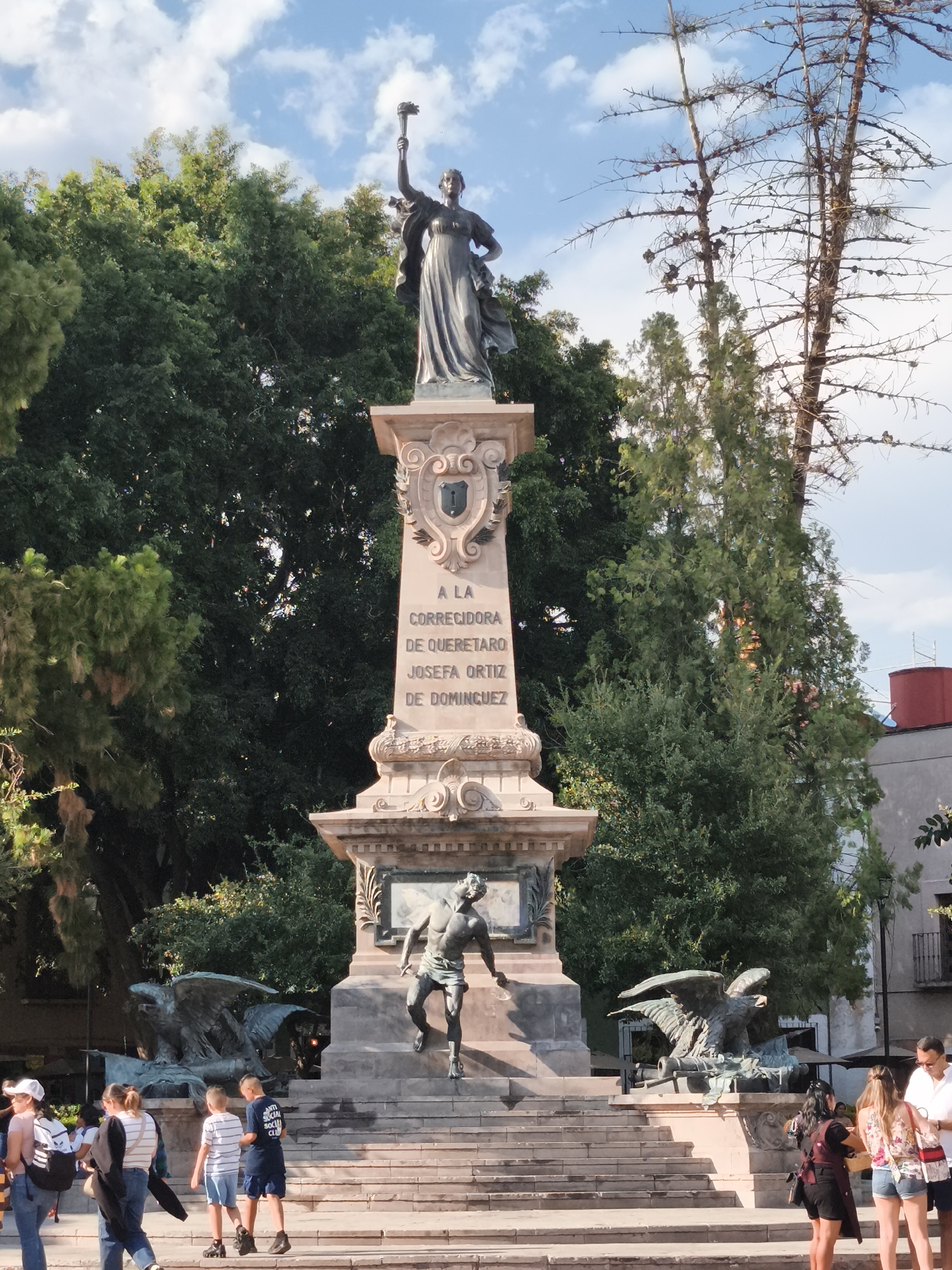 The image captures the Monument to Josefa Ortiz de Domínguez in Querétaro, Mexico. The monument features a tall stone structure with a statue of Josefa Ortiz de Domínguez holding a torch atop. Below, a bas-relief depicts a running figure. Bronze eagle sculptures adorn the base. A group of people are walking or standing on the steps leading up to the monument. The setting includes trees and a building in the background. The sky is partly cloudy.