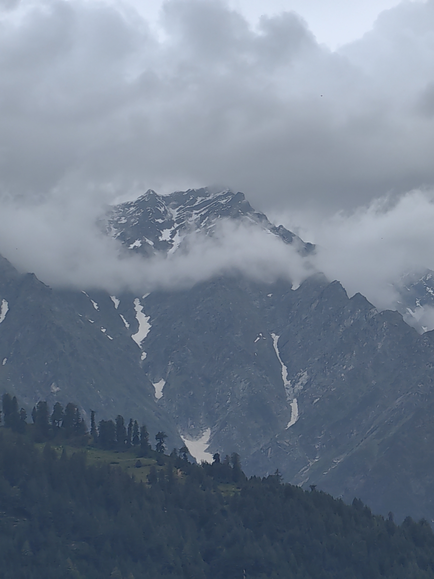 A mountain range under a cloudy sky is depicted. The mountains are rugged and steep, with some patches of snow visible on their slopes. Clouds partially obscure the upper peaks, creating a dramatic and atmospheric scene. In the foreground, a dense forest covers the lower slopes of the mountains, transitioning to a grassy area with scattered trees on a higher ridge. A small, isolated structure, possibly a hut or a small building, is visible on this grassy area. The overall impression is one of natural grandeur and remoteness. The location is specified as Vashisht, India, suggesting this is a view of the Himalayan foothills. There are no people or animals clearly visible in the image. The lighting is diffused, consistent with an overcast day.