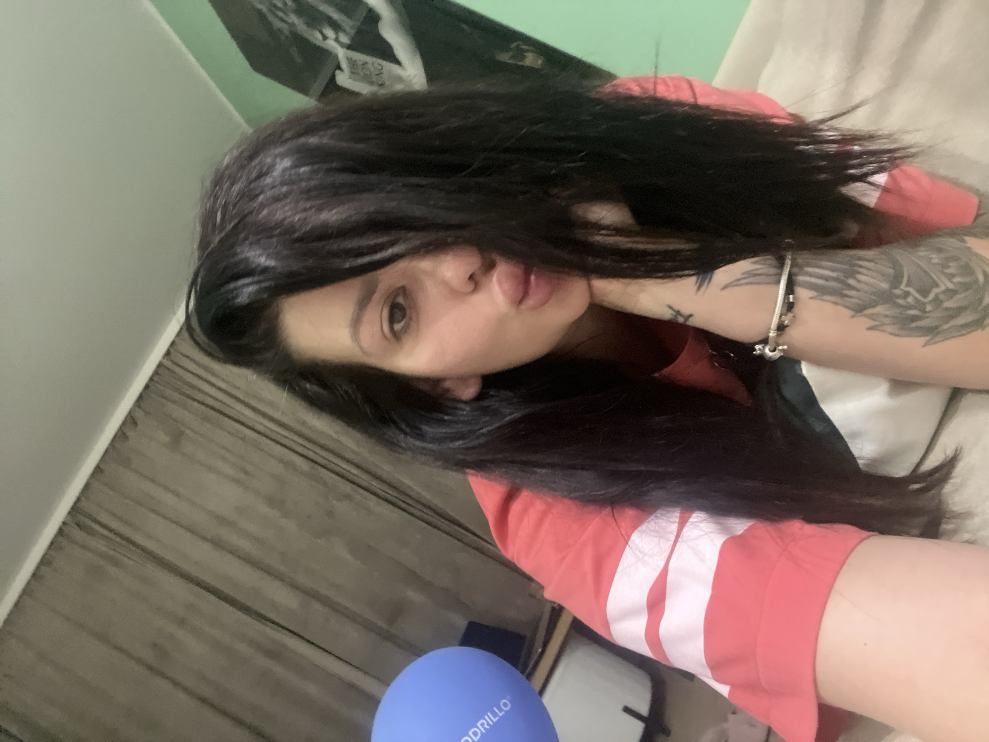 A selfie of a woman with long, dark hair covering part of her face, wearing a coral and white striped shirt, is taken indoors. She is leaning on her hand, with her lips pursed. A blue balloon with text is visible to her left. The background includes a muted brown curtain and a light green wall with a framed picture. The scene appears to be a residential interior. The image does not contain enough information to determine a specific location in the city of Plovdiv, Bulgaria.
