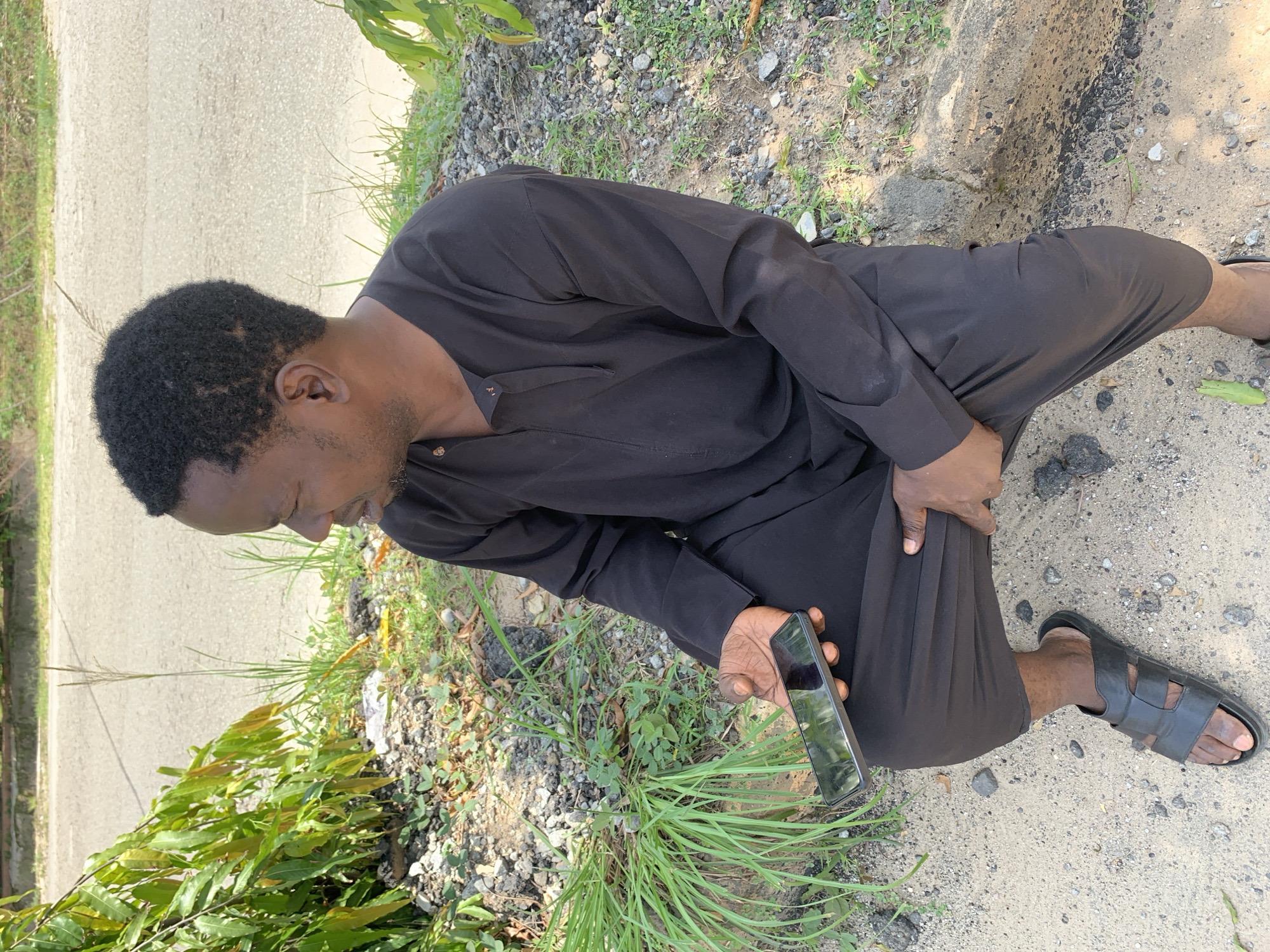A dark-skinned man with short black hair is sitting on the ground by the side of a paved road in Galadimawa Village, Nigeria. He is wearing a black long-sleeved traditional tunic and loose black pants. He is holding a black smartphone in his right hand, with the screen facing upwards. His left hand is resting on his left thigh, near the crotch of his pants. He is wearing black sandals. The ground around him is a mixture of sand, gravel, and some green vegetation. Trees and bushes are visible in the background, as well as the paved road. The lighting suggests it is daytime, with the sun casting some shadows.