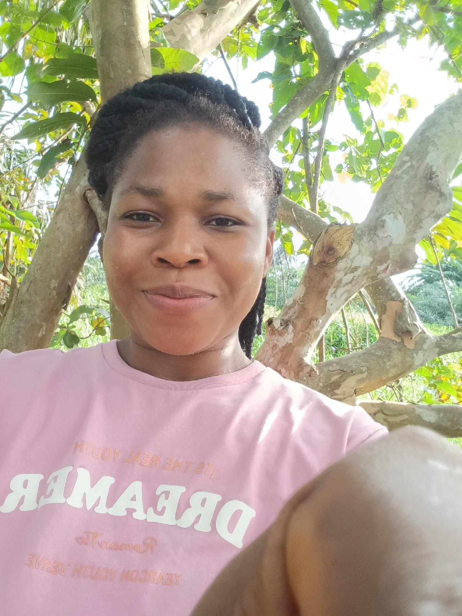 A woman with braided black hair, wearing a pink t-shirt with the word "DREAMER" in large white letters, smiles at the camera. She is positioned in front of a tree with light-colored bark and green leaves. The background is blurred with foliage and a bright sky, suggesting an outdoor setting. The image appears to be a selfie taken during daylight.