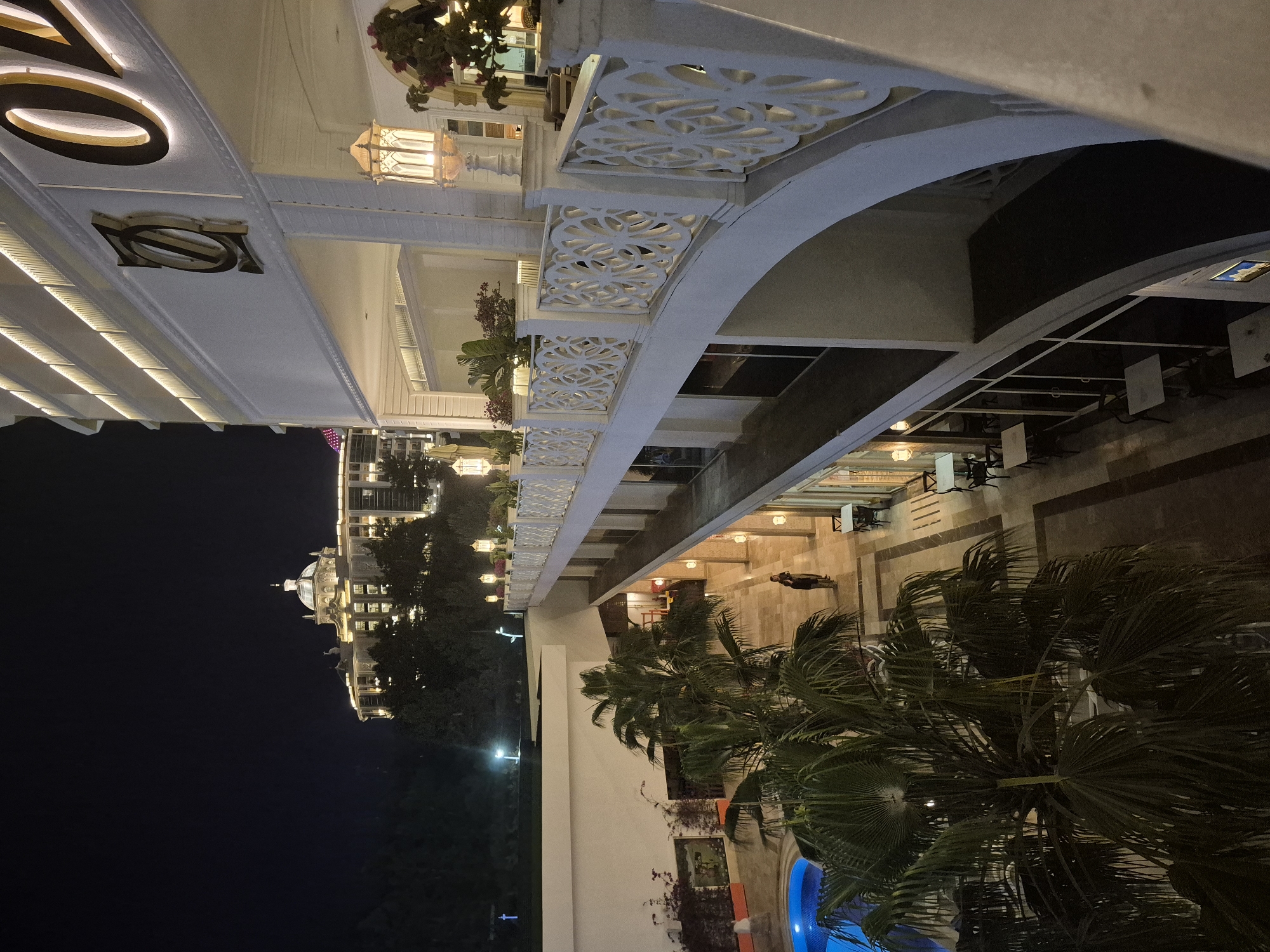 The image depicts a nighttime view of a luxury resort or hotel, possibly located in Alanya, Turkey. The scene is oriented vertically, showing the multi-level facade of a white building with ornate railings and architectural details. Palm trees are visible in the foreground, suggesting a tropical or coastal setting. Below the trees there is an edge of a swimming pool illuminated by blue light. A person is seen on the sidewalk between the building and the trees. The building is lit with warm lighting, highlighting its architectural features. In the background, another building with a dome can be seen. The night sky is dark, emphasizing the illuminated structures.