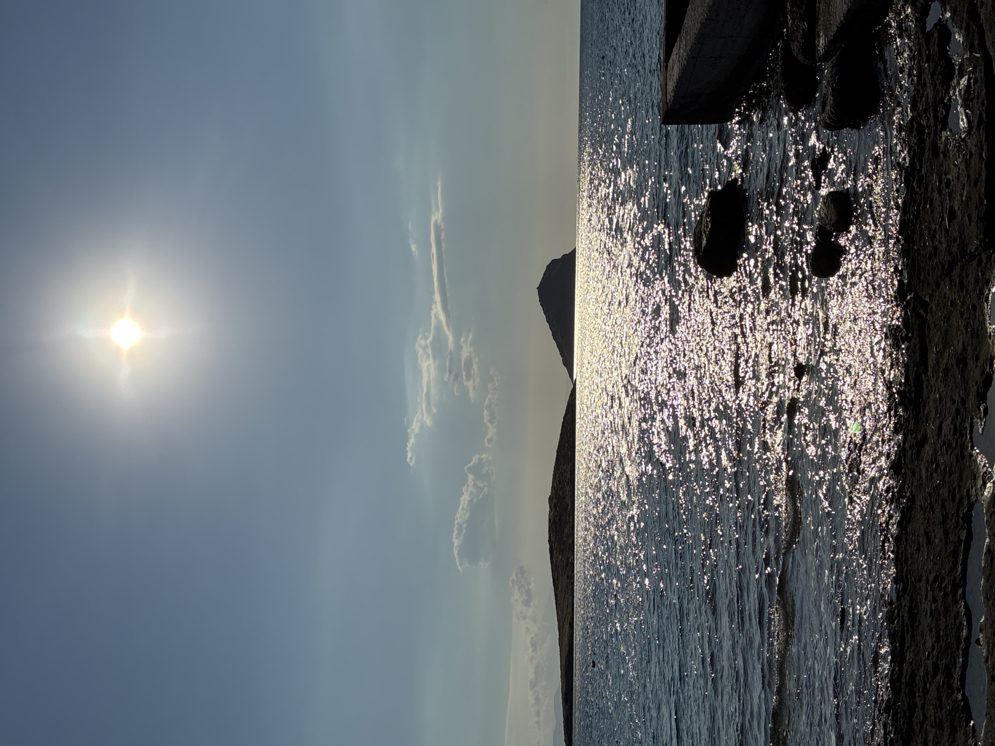 A sun glare over the Aegean Sea is captured in this vertical, outdoor, full shot of a seascape. The sun is positioned in the upper center of the frame, its rays illuminating the water with a brilliant sparkle. In the middle ground, two islands are visible in silhouette against the hazy horizon. The island on the left is low-lying and extends horizontally, while the island on the right is a more prominent, conical shape with a small structure at its peak. The foreground features a rocky shoreline with a concrete structure extending into the water on the right. The water is a deep blue, reflecting the sun's light in a shimmering pattern. The sky is a clear, pale blue with a few wispy clouds. The scene appears to be located in the vicinity of Limenas Markopoulou, Greece, given the characteristic island formations and the Aegean Sea. There are no people or animals visible in the image.