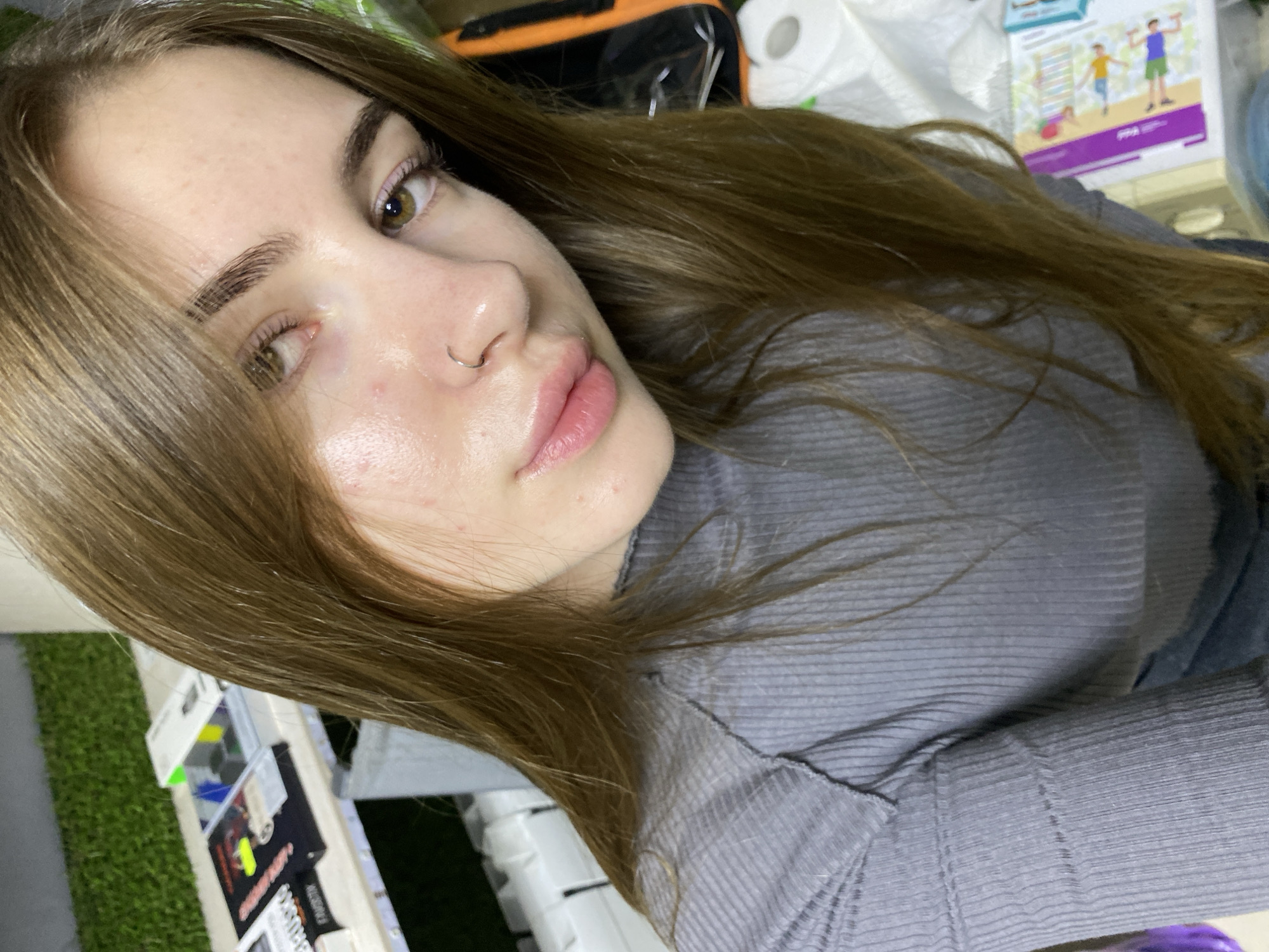 A young woman with long brown hair and a nose ring is shown in a close-up portrait, taken from a slightly upward angle. She is wearing a gray ribbed shirt. Her skin appears dewy, with some minor blemishes visible. Her eyes are green and she is looking towards the camera. The background is blurred, but suggests an indoor setting with shelves of books or similar items on the left and what appears to be toilet paper rolls and a bag on the right. There is no discernible connection to the city of Дьяково, Russia.