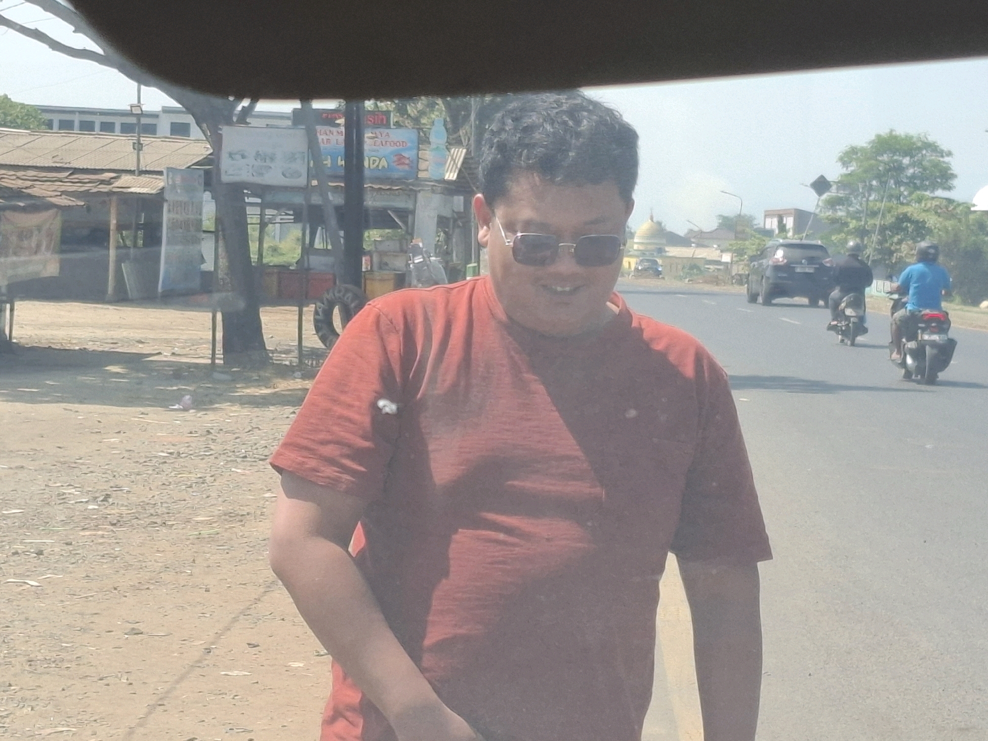 The image depicts a street scene in Babakan, Indonesia. In the foreground, a person wearing a red t-shirt and sunglasses is facing the camera. Behind them, a road with some traffic is visible, including two motorcycles and a black SUV. On the left side of the road, there are several small buildings, possibly market stalls or shops, with corrugated metal roofs. One sign above these structures reads "HAN MA YA CAFOD" and another with a picture of a bottle. The ground is dusty and uneven, suggesting a rural or semi-developed area. The sky is bright and hazy, indicating a sunny day. The overall atmosphere appears to be casual and everyday.