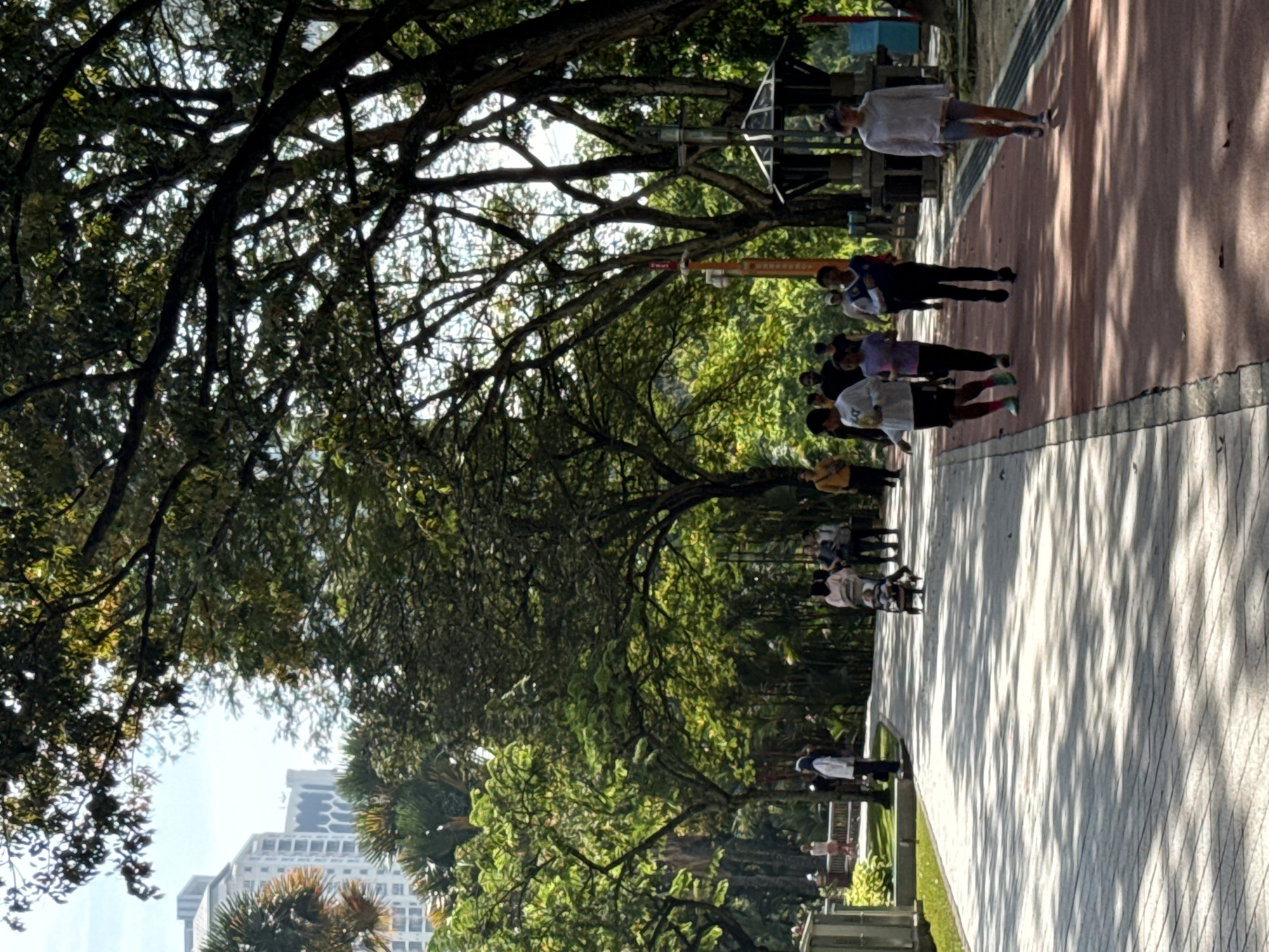 The image depicts a public park in Kuala Lumpur, Malaysia. A group of people, mostly young, are walking along a tiled path. The park is densely populated with trees that provide shade. In the background, modern buildings are visible. The overall scene conveys a sense of urban nature and casual activity.