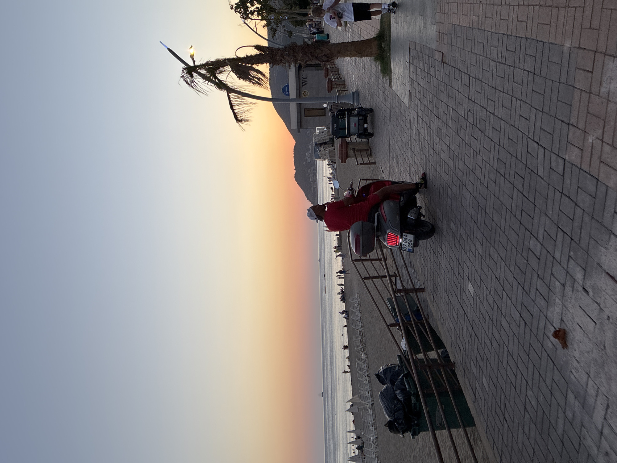 An outdoor, eye-level, vertical shot shows a paved walkway next to a beach at sunset in Alanya, Turkey. The sky is a gradient of pale blue to orange. A man in a red shirt and shorts rides a red scooter on the walkway. The scooter has a license plate that reads "07 BKA 358." To the left of the walkway is a sandy beach with rows of empty lounge chairs. There are also large green trash bins on the beach. Further out in the sea, a boat is visible. Behind the walkway, there is a palm tree and a building with a "WC" sign. A young boy in a white t-shirt and black shorts walks away from the viewer on the walkway, holding a drink. Another person is walking in the background. The walkway is paved with rectangular bricks.