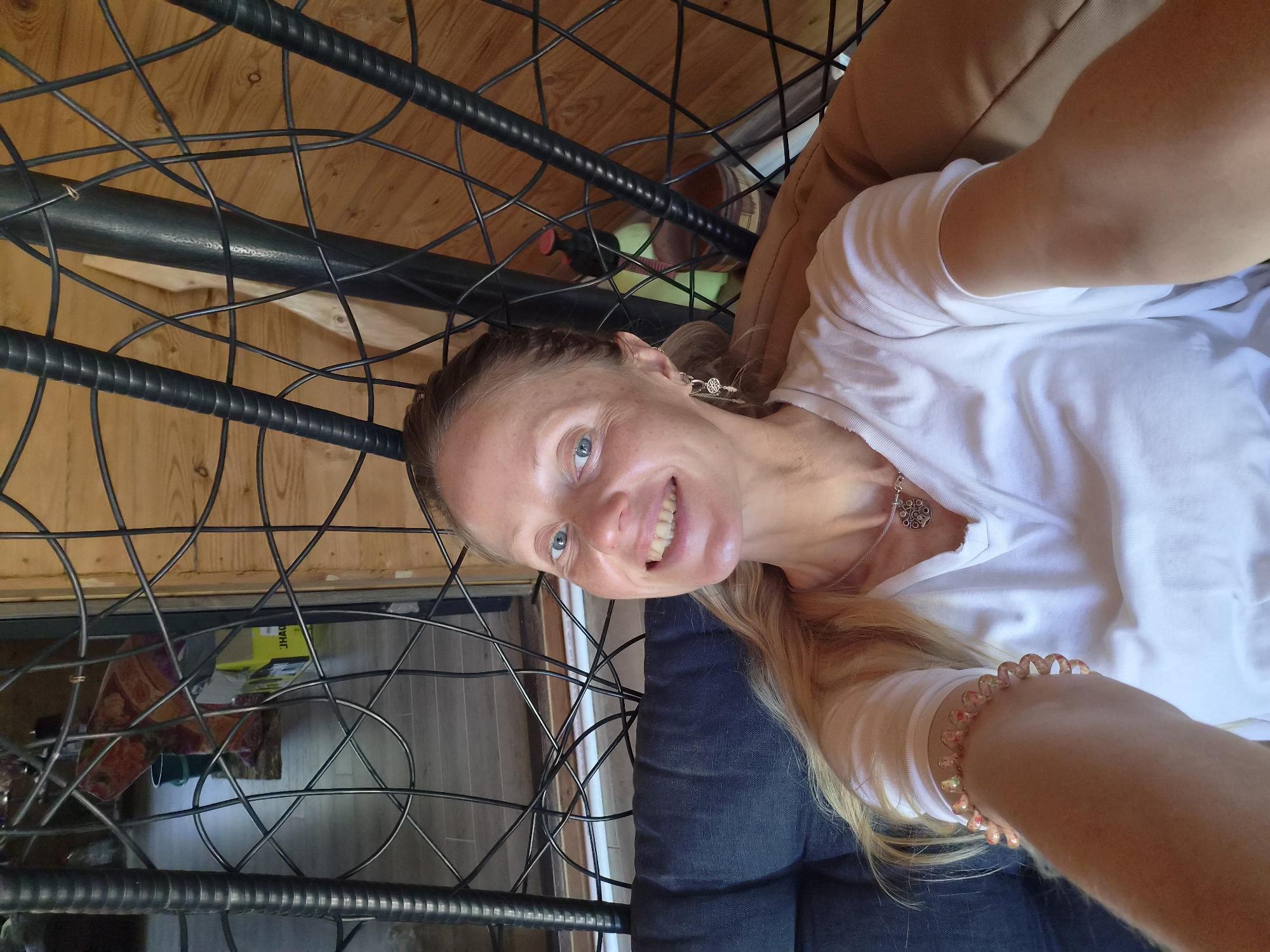 An adult female is depicted in a selfie, seated in a dark-framed, woven-style swing chair with dark grey and tan cushions, in an indoor setting located in Motyakovo, Russia. She has fair skin, blue eyes, and light blonde hair pulled back. Her expression is a broad smile, revealing her teeth. She wears a white V-neck t-shirt, a silver-colored necklace with an ornate circular pendant, and multiple earrings on her right ear, including a small hoop and dangle charms resembling a tree of life and a feather. On her left wrist, she wears a light-colored spiral hair tie or bracelet. The background behind her features light-toned horizontal wooden paneling walls and grey-toned tiled flooring. Visible through the chair's frame are various objects: a dark green bucket, a patterned fabric in red and purple tones, a yellow box with "JHAC" visible, a light green plastic bottle with a red pump top, and a dark brown container. A light wooden board is also partially visible in the upper right background.