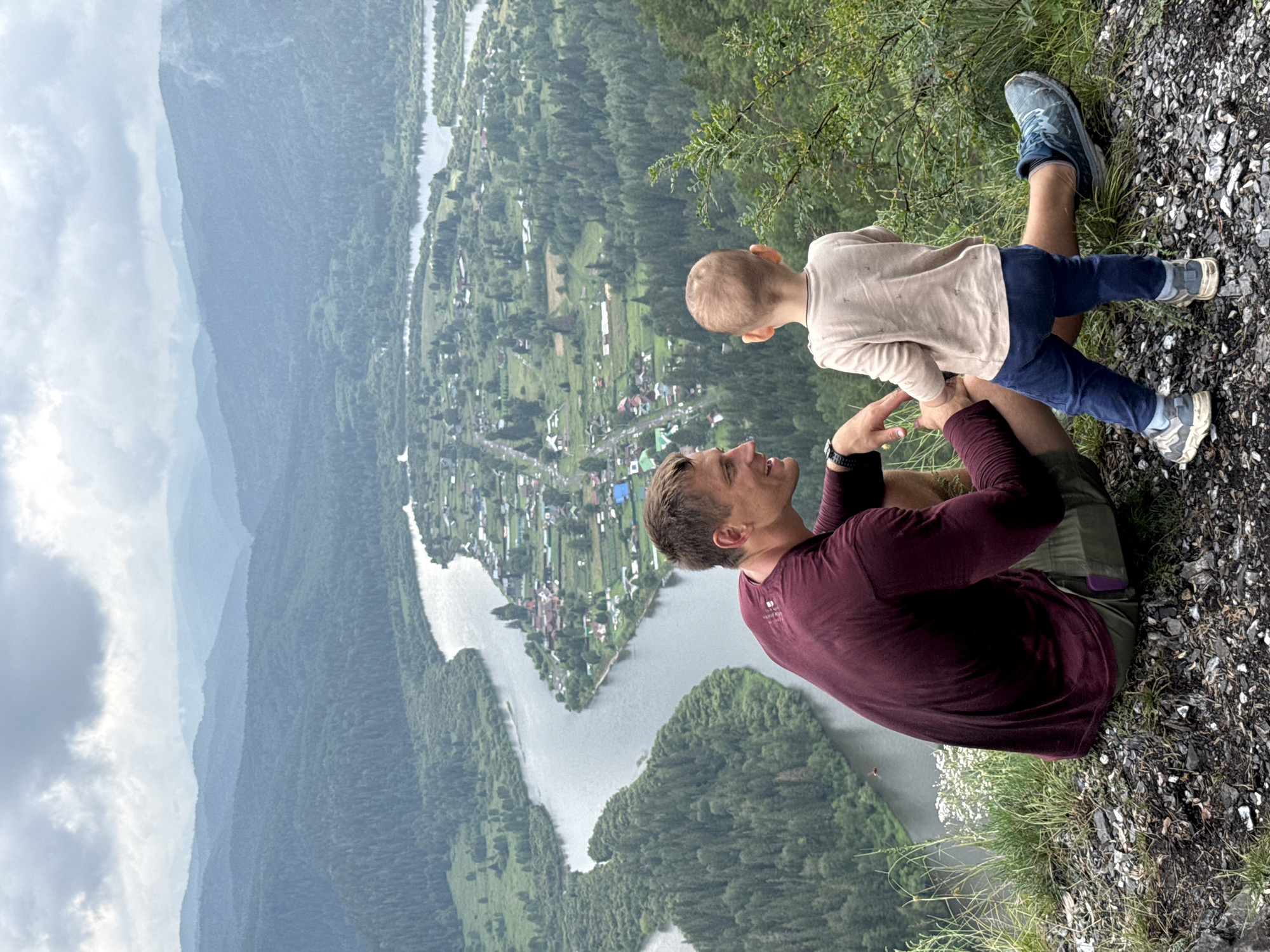 A man and a young child are seated on a rocky outcrop overlooking a valley. The man, wearing a maroon long-sleeved shirt and olive shorts, is looking at the child. The child, wearing a light-colored t-shirt and dark blue pants, is facing away from the man, looking out at the valley. The valley features a winding river, a small village with numerous buildings, and dense green forests covering rolling hills that extend into the distance. The sky is overcast with clouds. The location is the viewpoint overlooking the city of Urman, Russia.