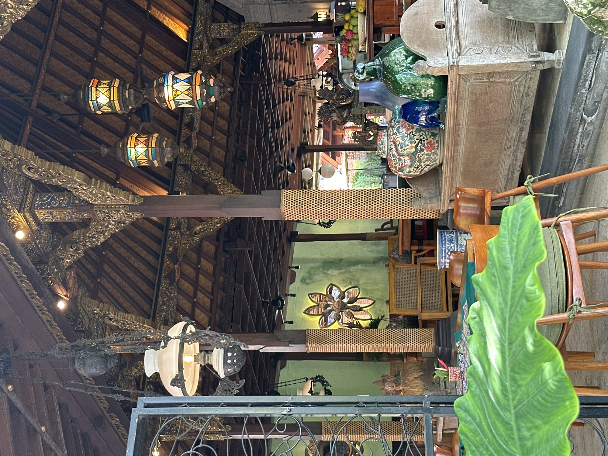 This is an interior shot of a restaurant in the Ubud District, Indonesia. The scene is dimly lit and features traditional Balinese decor. In the foreground, a large green leaf partially obscures a wooden chair with a green cushion. In the midground, there is a wooden chest adorned with decorative carvings, topped with a variety of vases and jars, including a large green glass bottle and a colorful ceramic vase. Behind the chest, various wooden chairs and tables are visible, along with decorative elements such as hanging lanterns and a flower-shaped mirror on a light green wall. The ceiling is made of dark wood with intricate golden carvings and features multiple hanging lamps, including traditional kerosene lamps and multi-colored lanterns. A statue of Buddha is also visible in the background. The overall atmosphere suggests a cultural and artistic dining establishment.