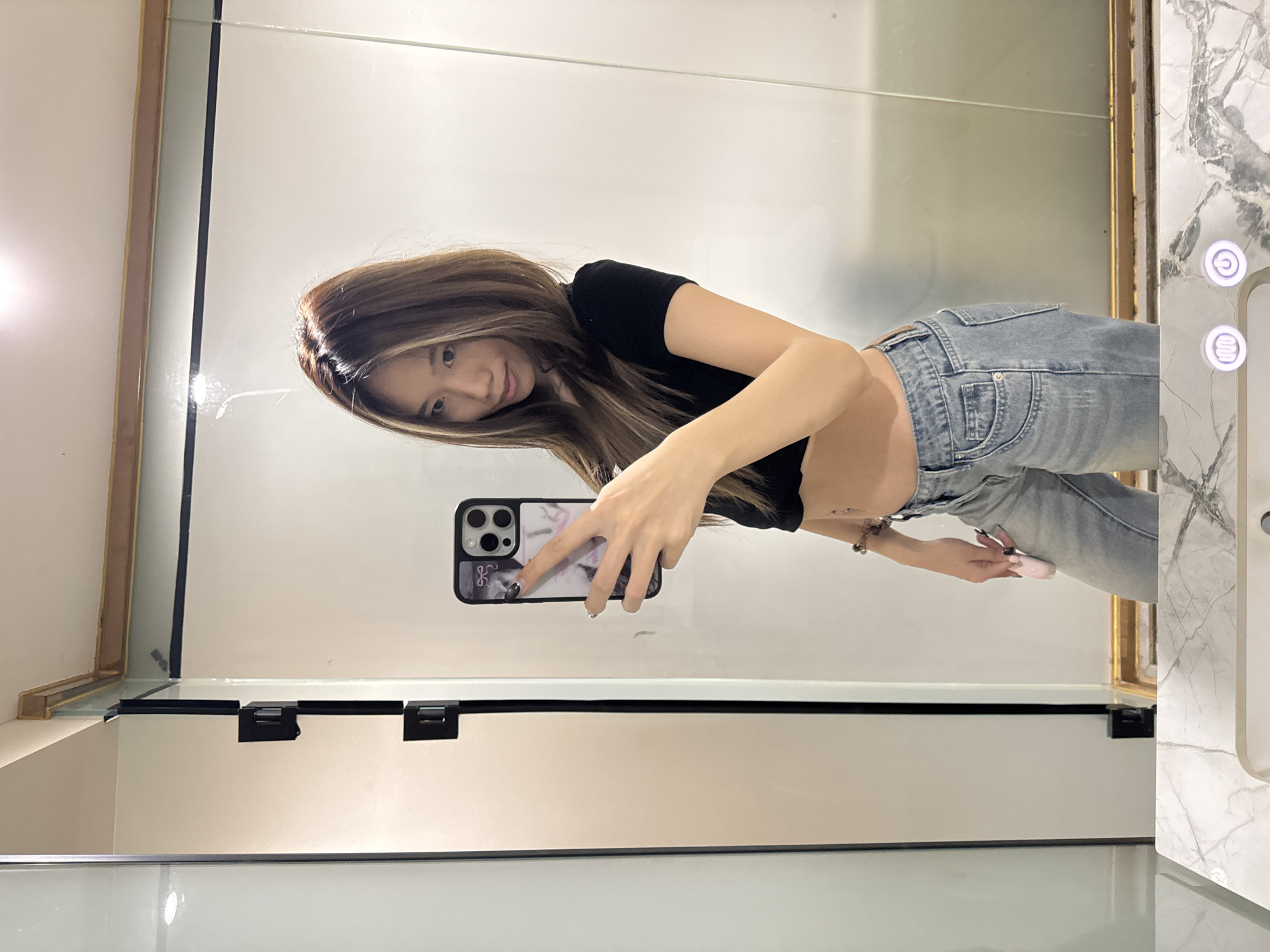 A young woman is taking a selfie in a bathroom. She is wearing a black crop top and light blue jeans. She is holding a smartphone with a patterned case. The bathroom has a large mirror, marble countertops, and white cabinets. There is no information to determine if this photo was taken in the city of Yuzhong District, China.