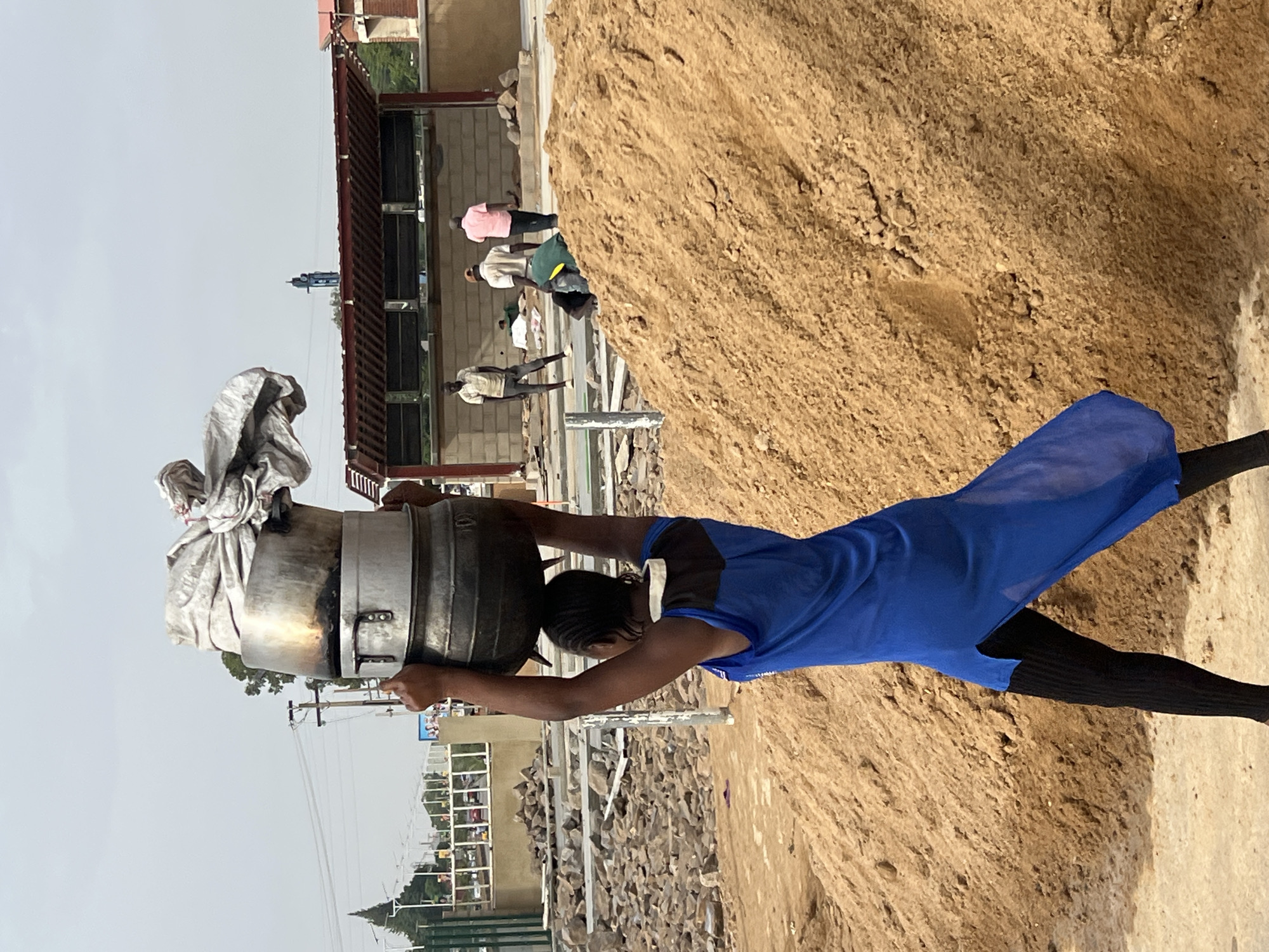 In Barakin Lerawi, Nigeria, a person in a blue dress and black leggings carries a large, covered metal pot on their head near a sand pile. Other people are present near a building under construction in the background.